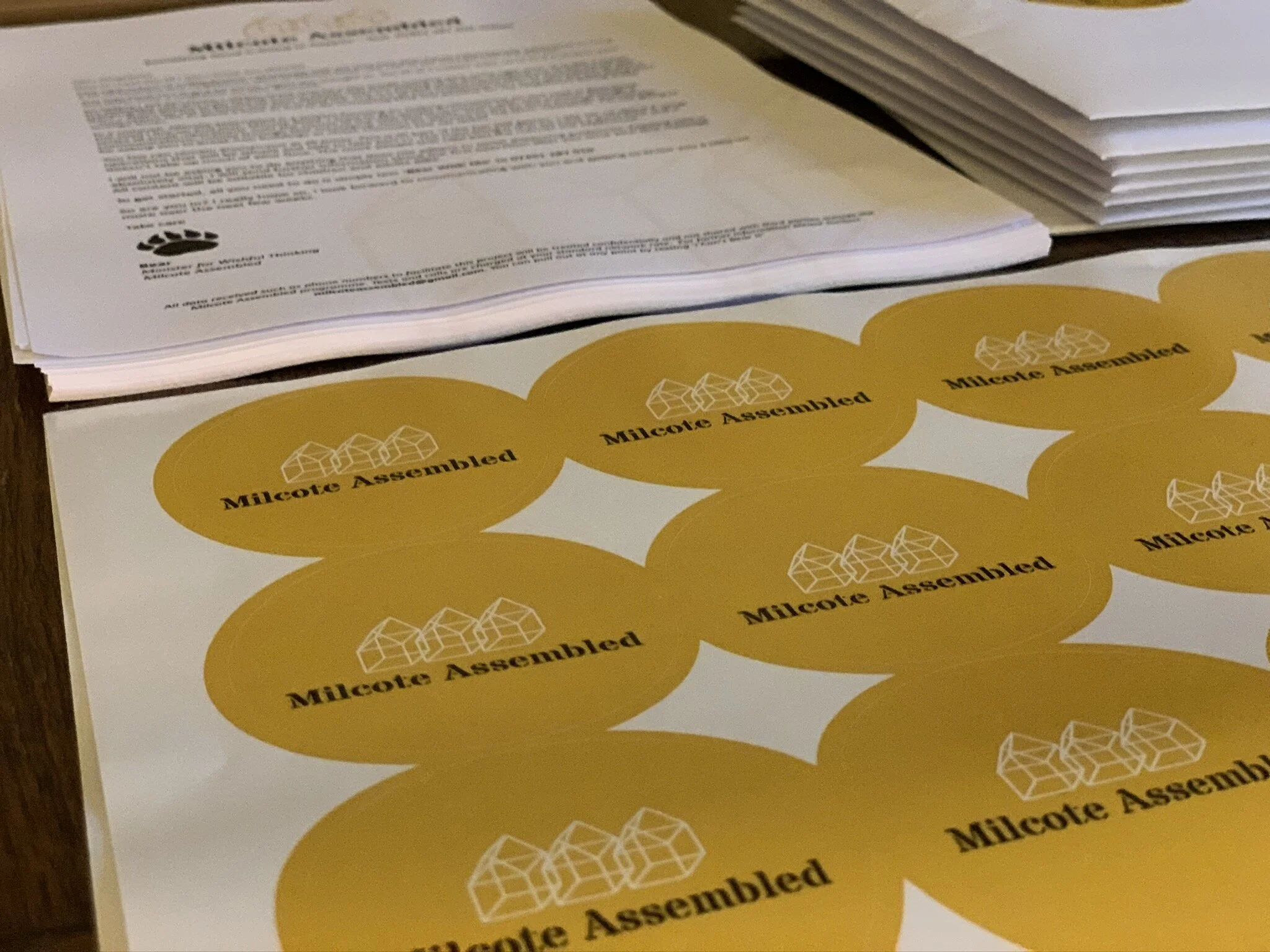 A stack of papers and a sheet of circular yellow stickers with the text 'Milcotê Assembled' and a geometric logo.