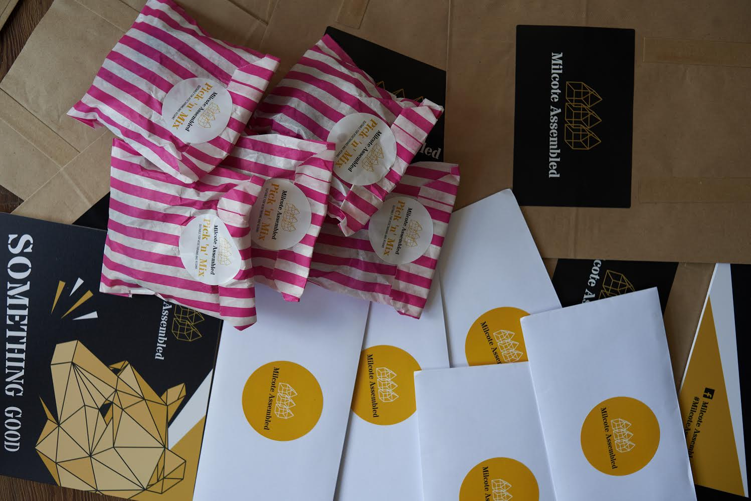 Multiple pink and white striped packages with circular labels, some open, on a wooden table along with black and white envelopes and folders with gold geometric logos and text for Milletke Assembled.