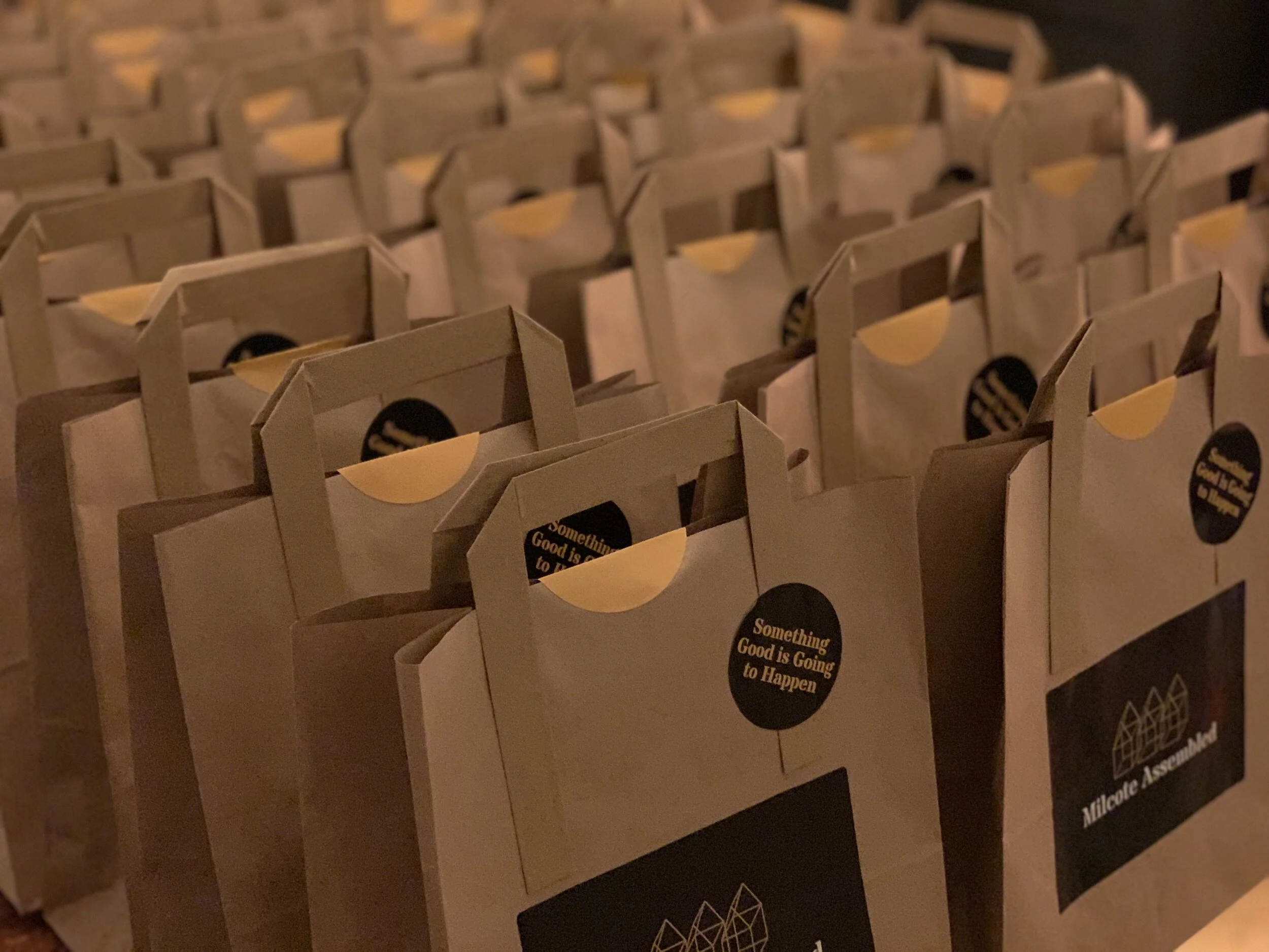 A close-up of multiple paper gift bags with handles, featuring a black circular sticker that reads 'Something Good is Going to Happen' in yellow text.