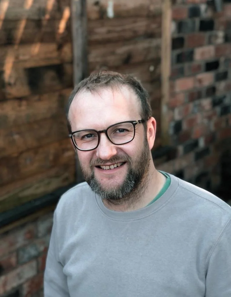 A smiling man with glasses and a beard standing outdoors in front of a wooden fence.