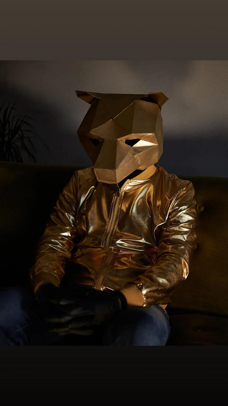 Person sitting on a couch wearing a shiny, metallic gold jacket and black gloves, with a geometric golden bear mask over their face, in a dimly lit room.