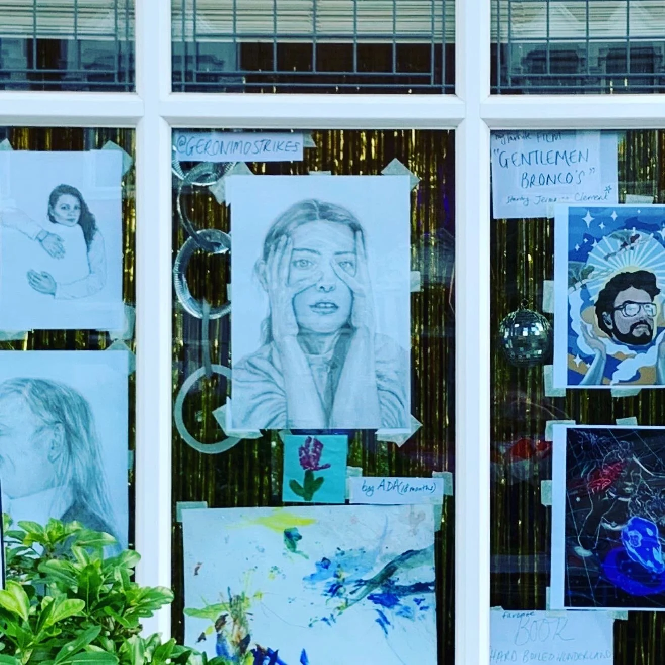 Window display with various artwork including a pencil sketch of a woman holding her hands to her face, a drawing of a girl with long hair, and other colorful art pieces. Handwritten notes and signs are also visible.