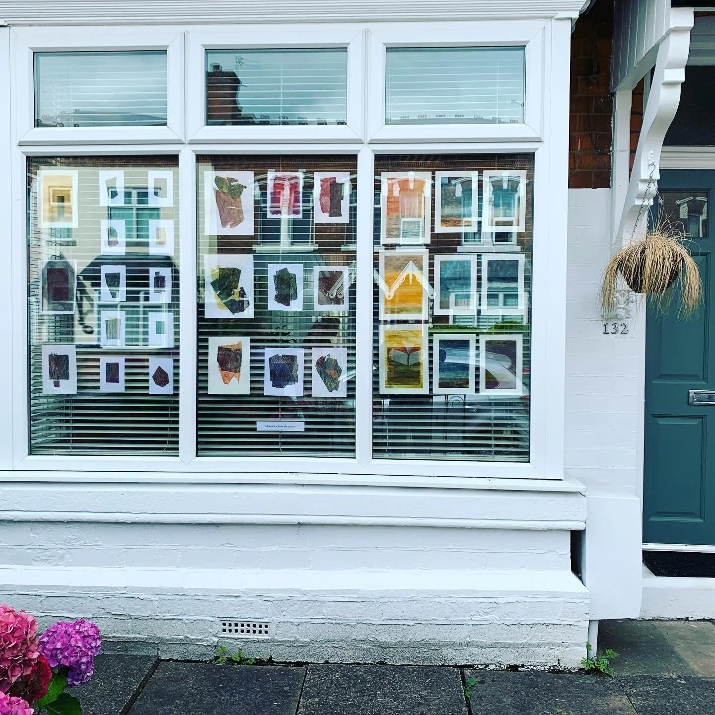 Display window with artwork of colorful rocks, framed and hung on paper, outside a house with a green door and pink flowers in the foreground.