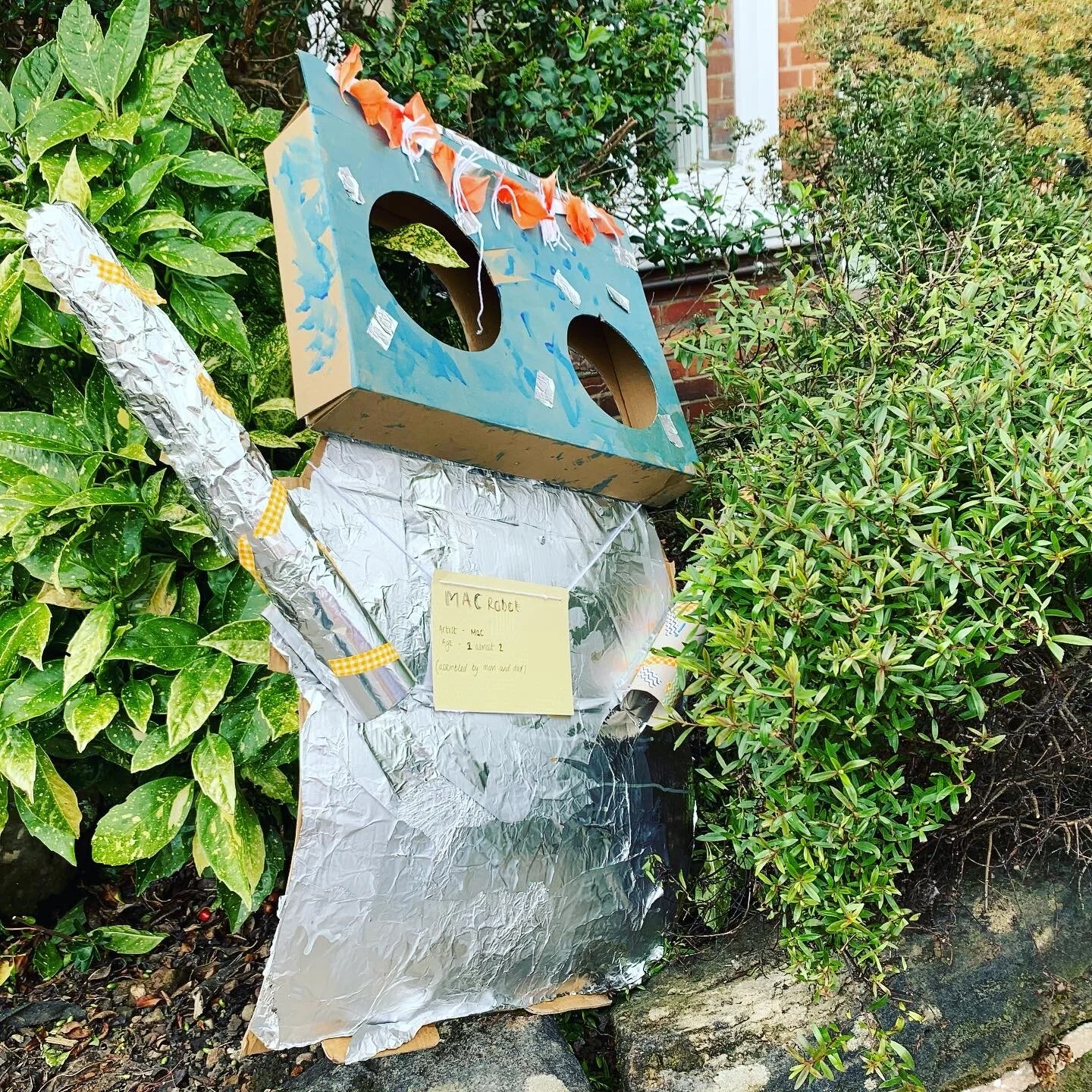 A robot made from a cardboard box, painted to resemble a robot with a blue face and orange accents. The body is covered in aluminum foil and decorated with taped embellishments. The robot is placed outdoors among green bushes in front of a brick hous