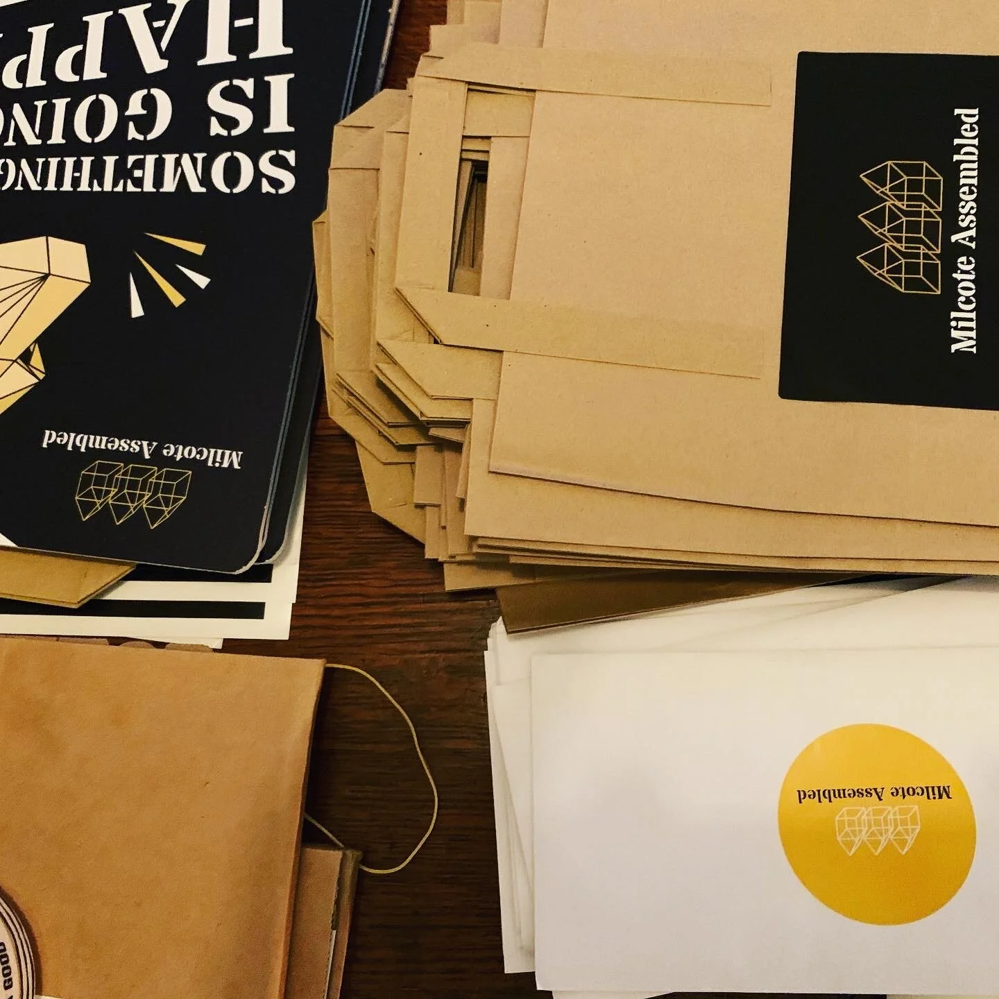 Stacks of paper folders and brochures on a wooden table, some labeled with 'Milcoote Assembled' and geometric logo, in black, white, and yellow colors.
