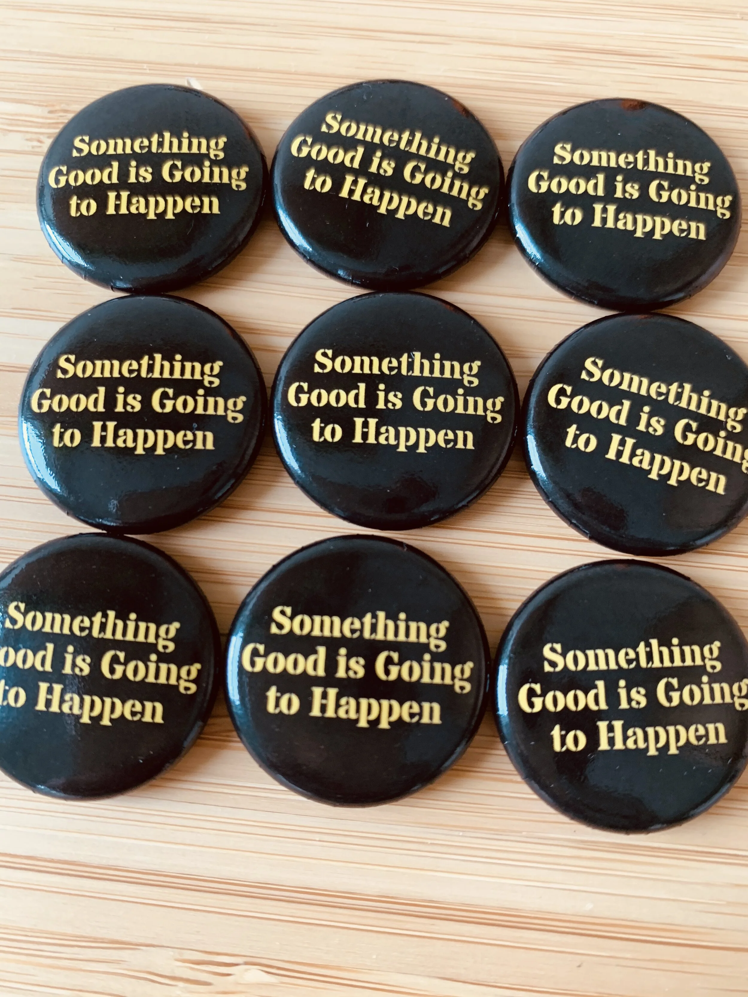 Six black circular buttons with yellow text that reads, 'Something Good is Going to Happen,' arranged on a light wooden surface.
