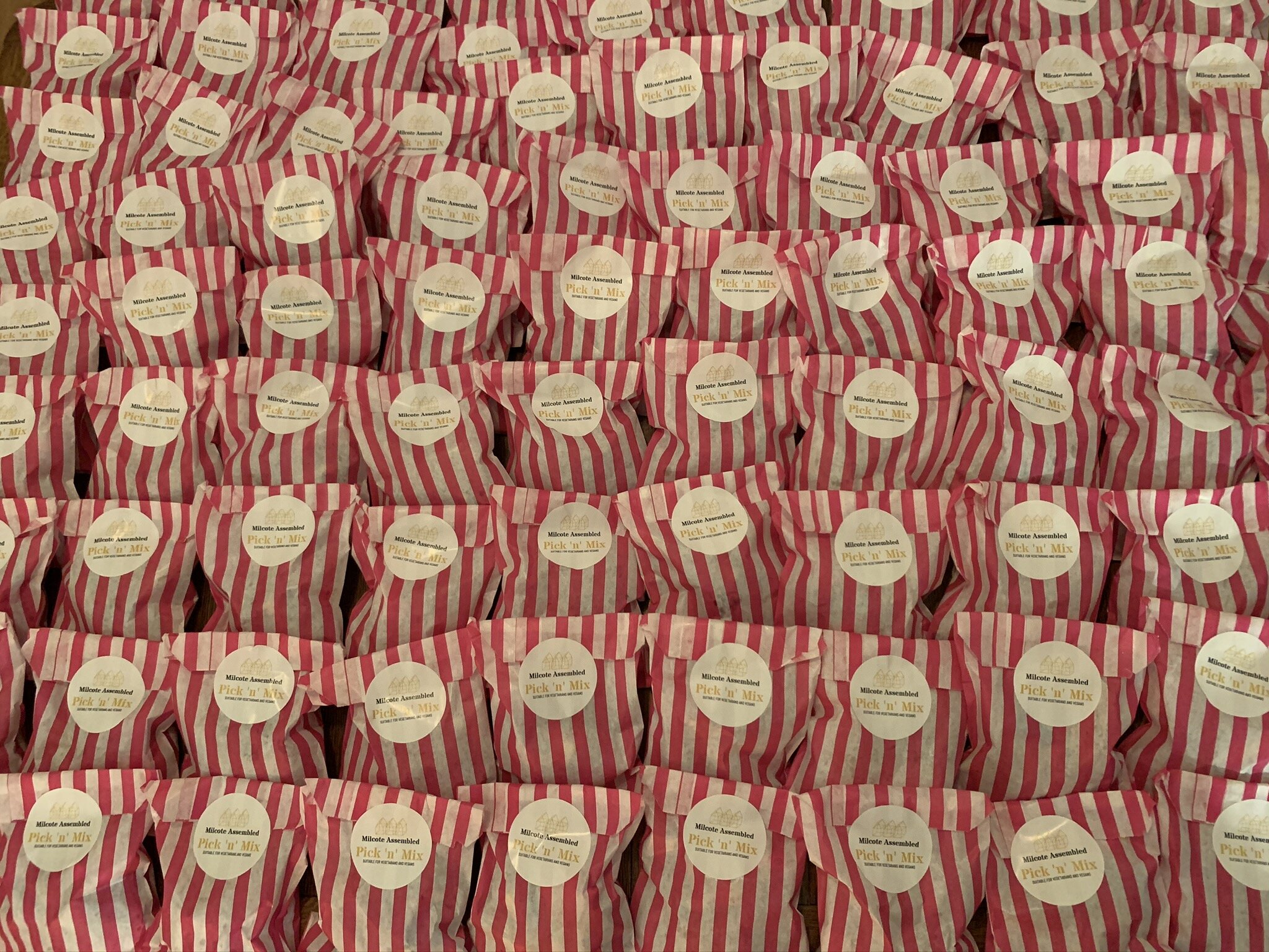 Multiple red and white striped paper bags with white circular labels that read 'Milcote Assembled Pick 'n' Mix, suitable for vegetarians and vegans.'