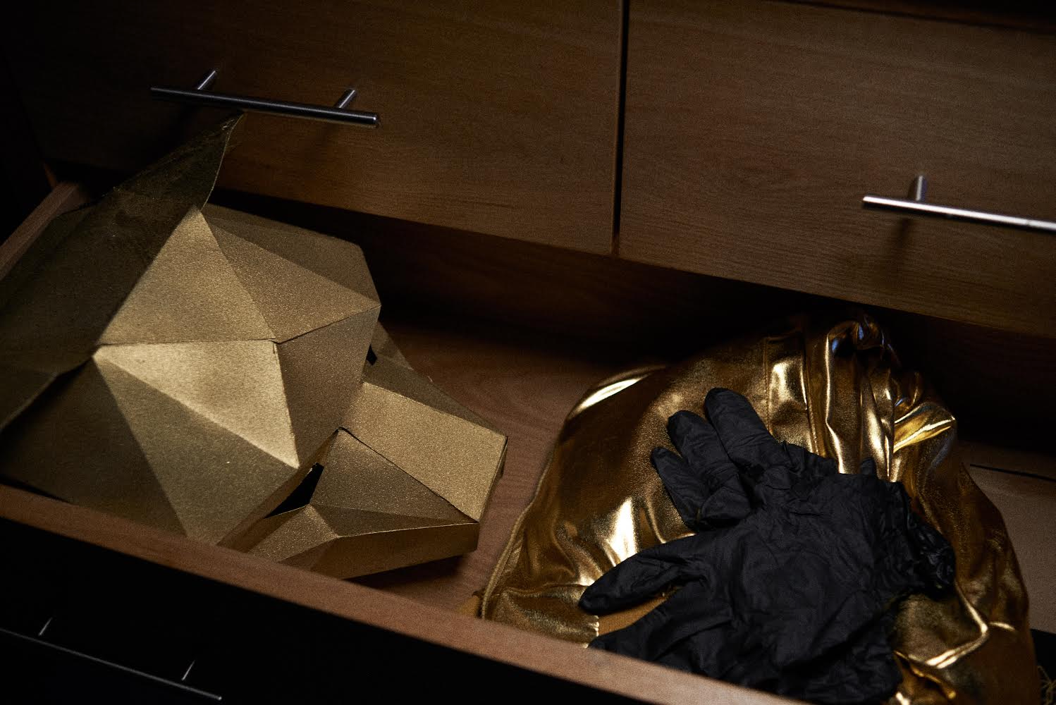 Open drawer containing a black glove and a gold metallic mask, with a gold geometric object on the left.