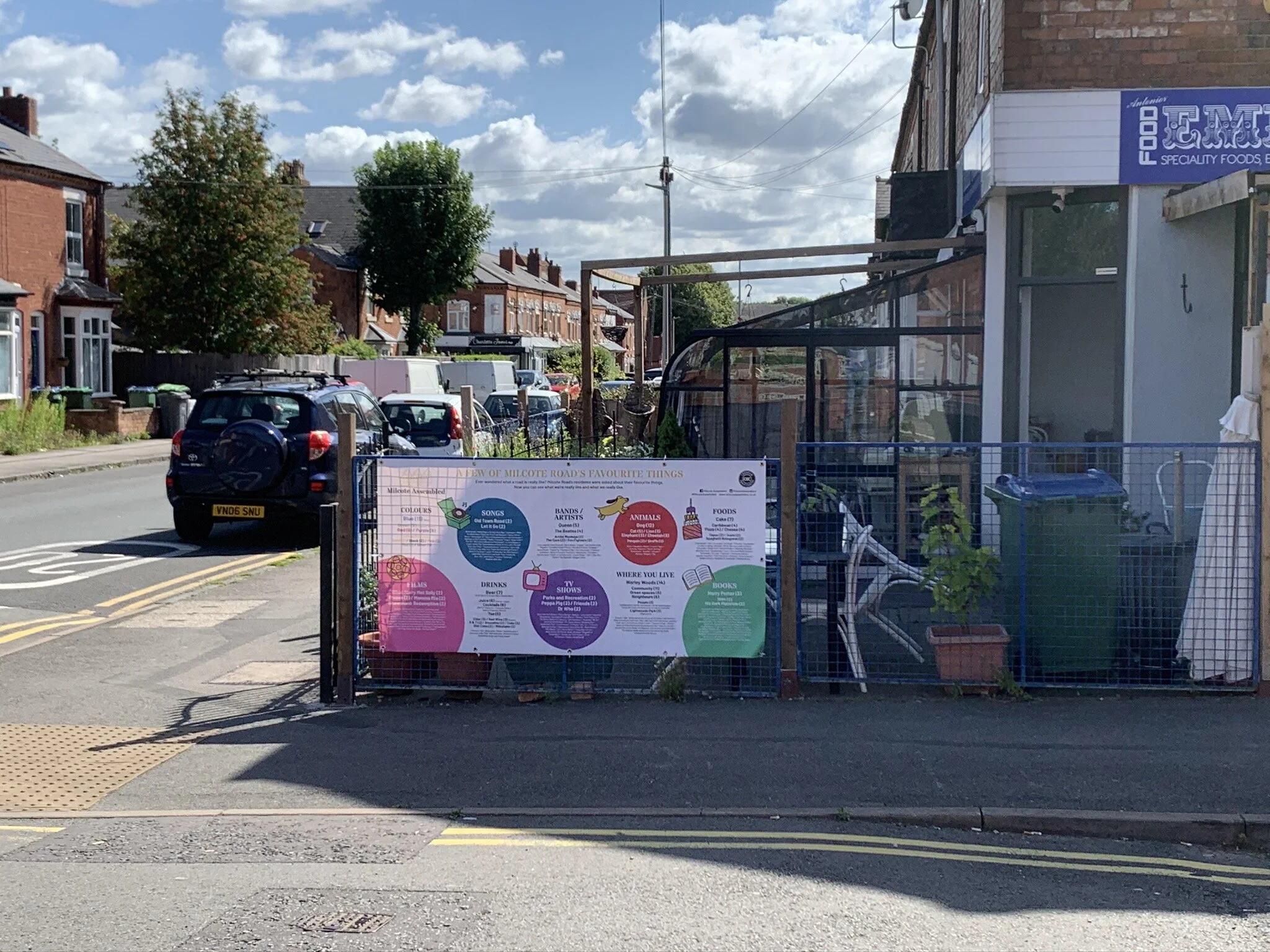 Street view showing parked cars, a building with a purple sign, trees, and a poster on a fence advertising community activities.