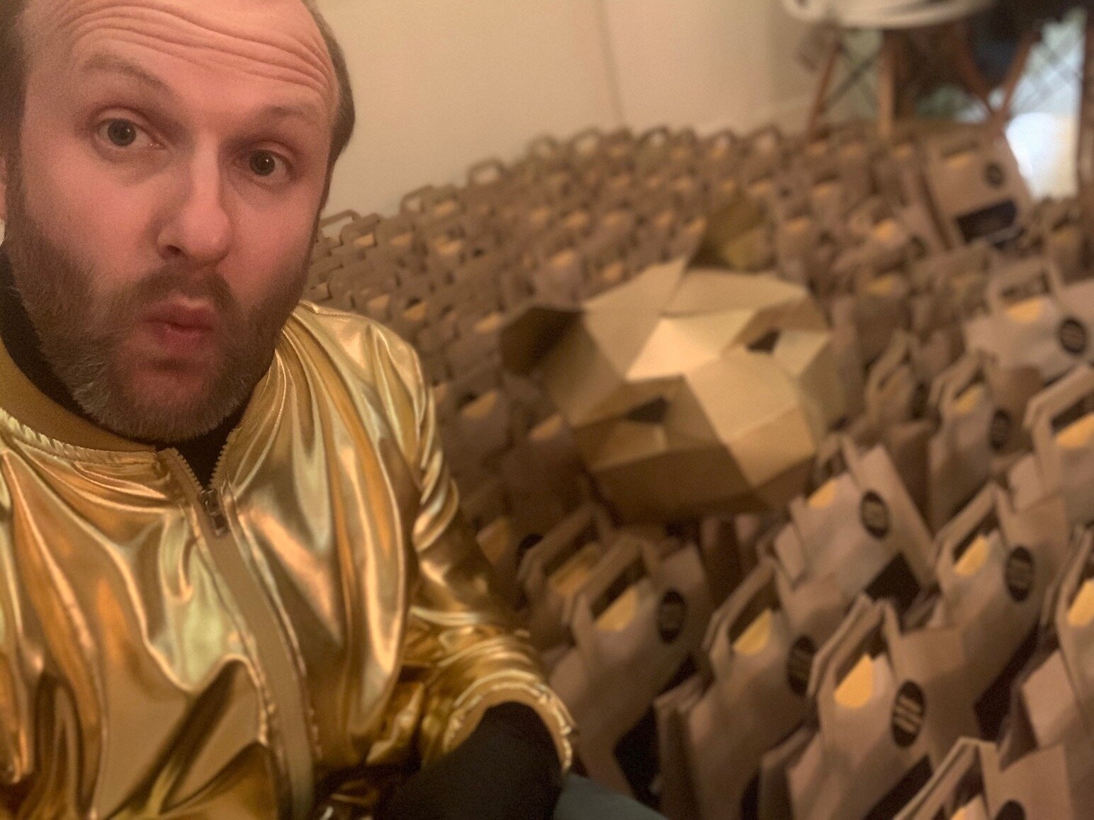A man with a beard wearing a shiny gold jacket taking a selfie in front of a large wooden sculpture of a sports car surrounded by wooden boxes.
