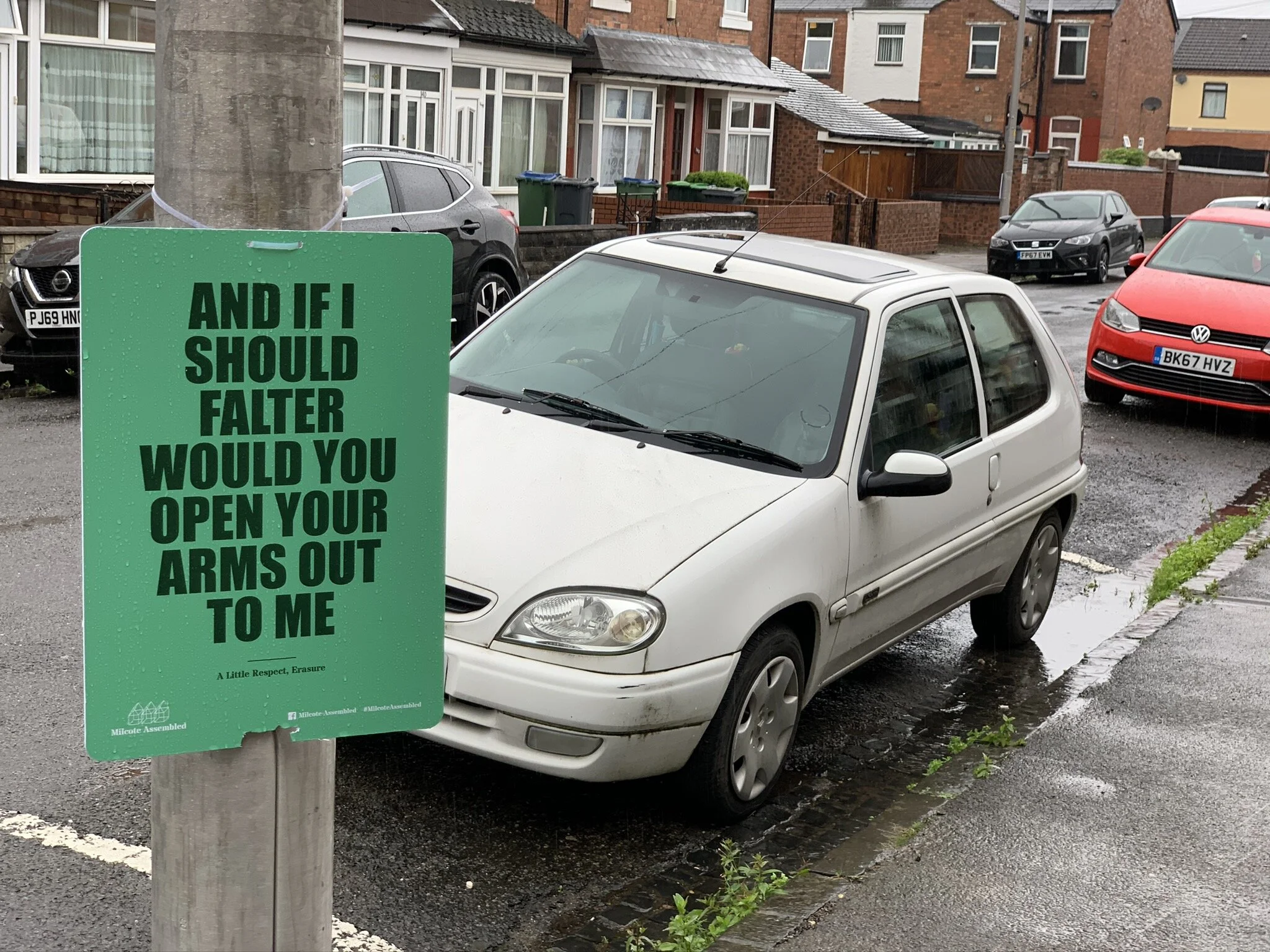 A parking lot with several cars parked in front of brick houses. A green sign with black text is attached to a wooden post, partially obscuring a white car in the foreground. The sign reads: "AND IF I SHOULD FALTER WOULD YOU OPEN YOUR ARMS OUT TO ME.