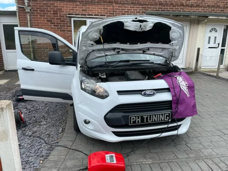A white Ford van with the hood open in a driveway, displaying "PH Tuning" on the front plate, and a purple cover on the front. A small red diagnostic device is placed on the ground next to it.