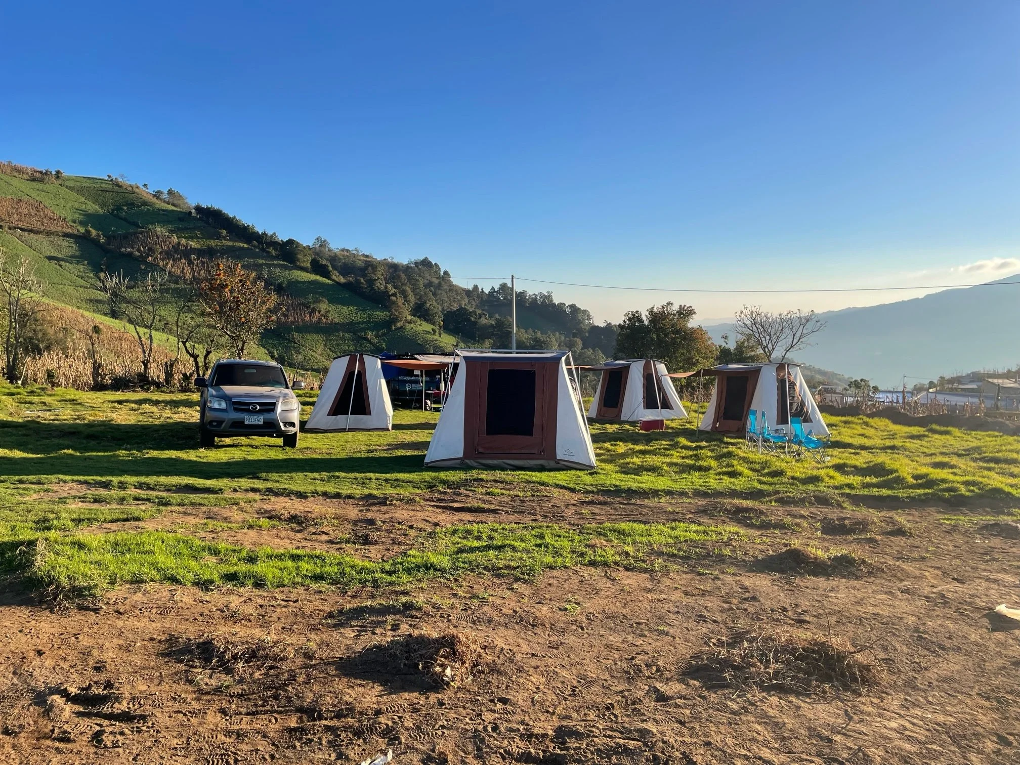 A camping scene with multiple tents and a parked SUV on grassy ground, set against green hills and a clear blue sky in the background.