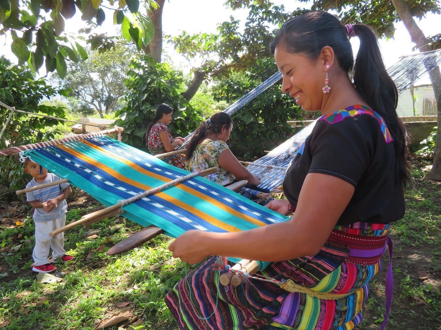 Women and children weaving colorful fabric on traditional looms outdoors in a green, shaded area in Lake Atitlan Guatemala