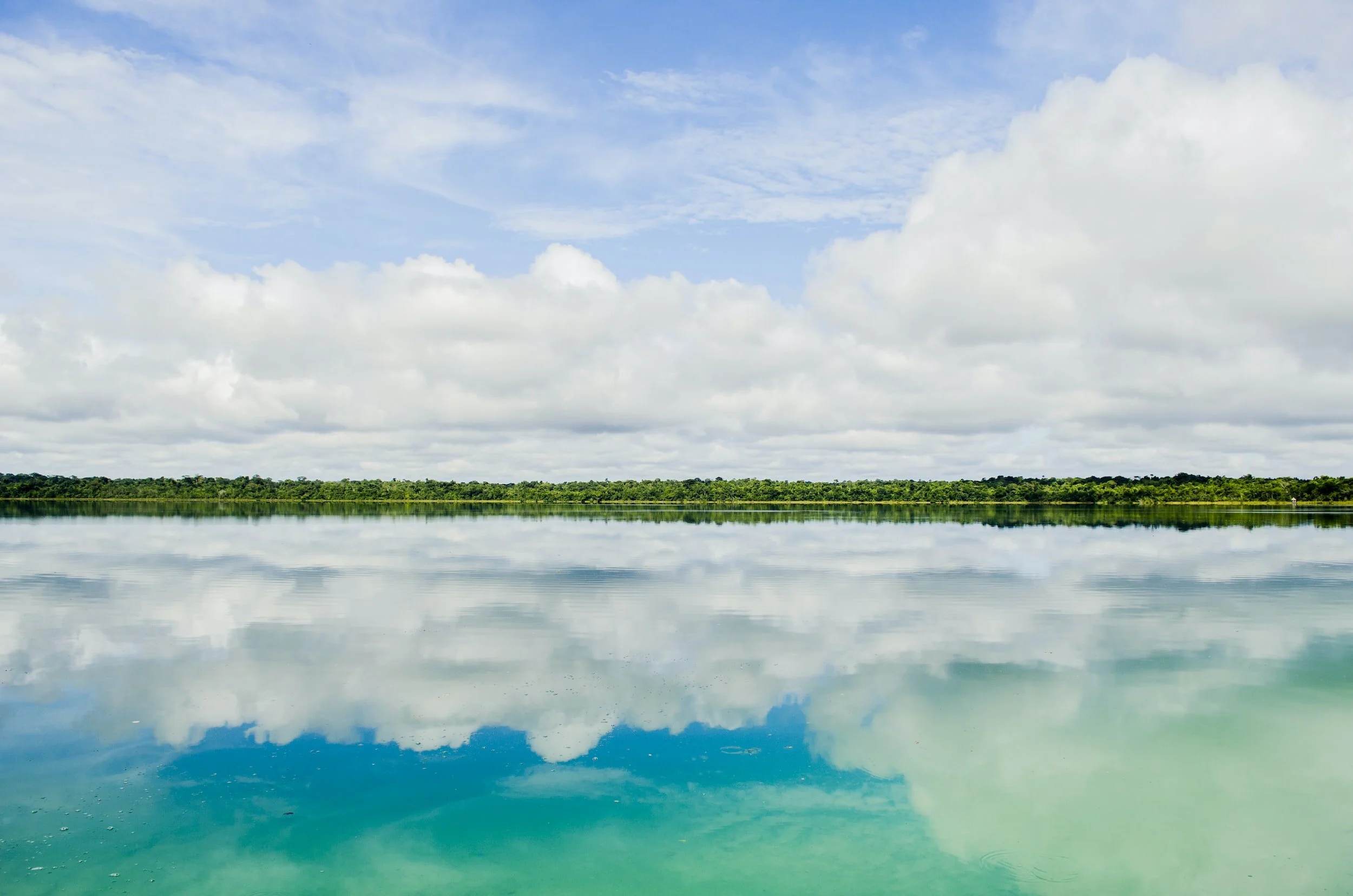 Calm lake reflecting a cloudy blue sky with a distant green shoreline.