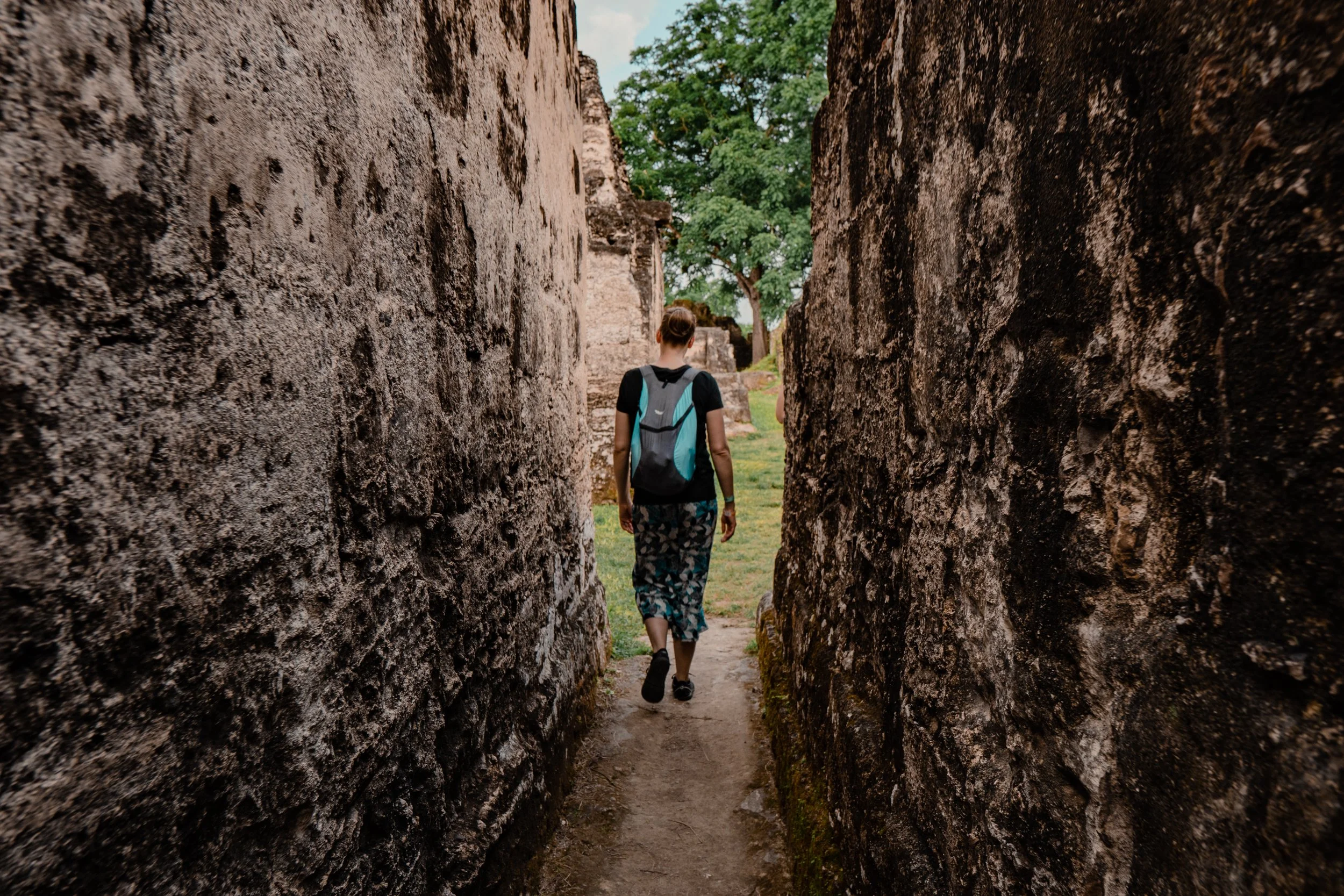 Person walking through a narrow passageway in Tikal, Guatemala carrying a backpack and wearing casual clothes and sneakers, with trees visible in the background.