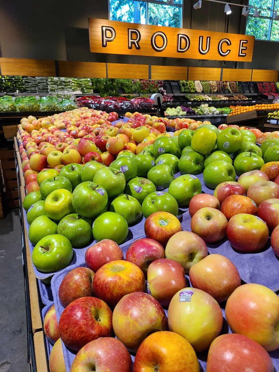 Fresh apples displayed in a produce section of a grocery store, with a sign above reading 'PRODUCE' and various other fruits and vegetables in the background.