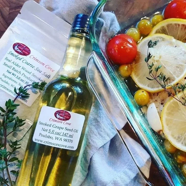 A bottle of smoked grape seed oil, a bag of smoked coarse sea salt, and a glass dish with cherry tomatoes, lemon slices, garlic, and sprigs of thyme.