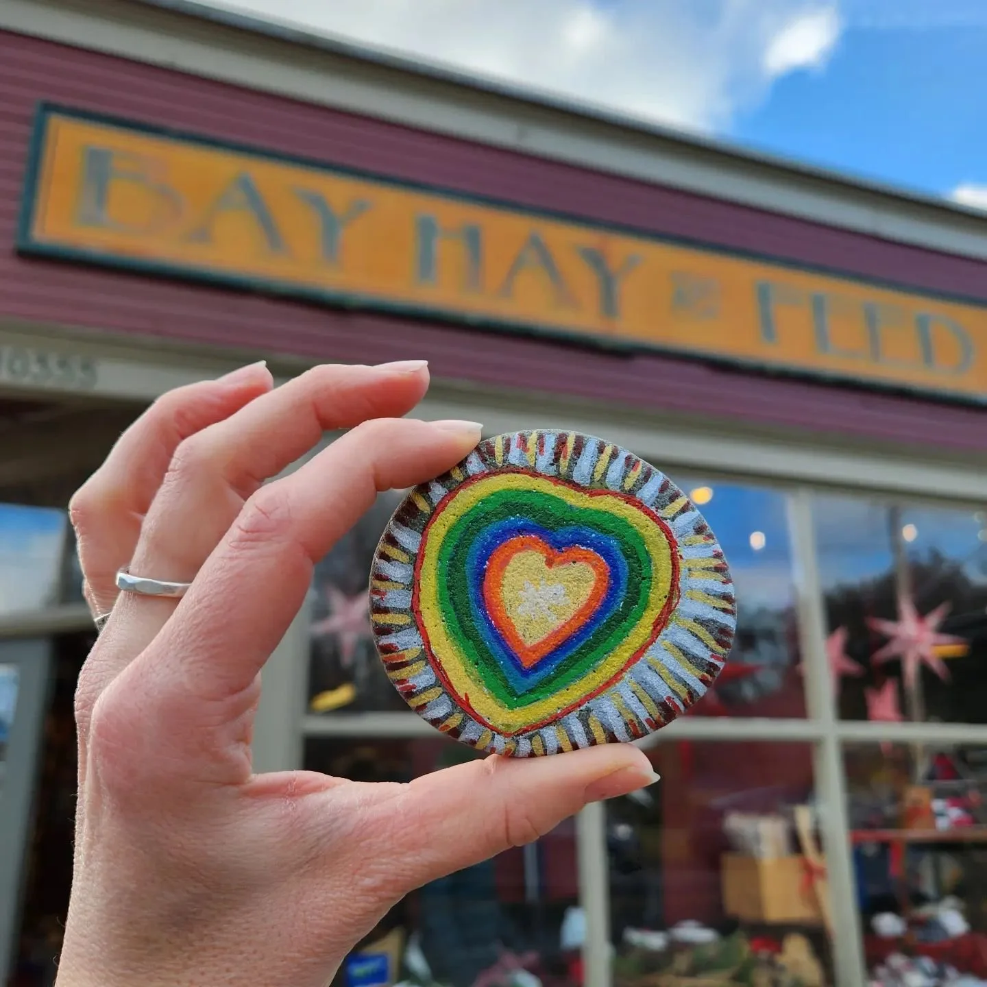 A hand holding a painted, rainbow-colored stone with a heart design in front of a storefront reading "Day Hay & Ltd." with windows showing decorations inside.