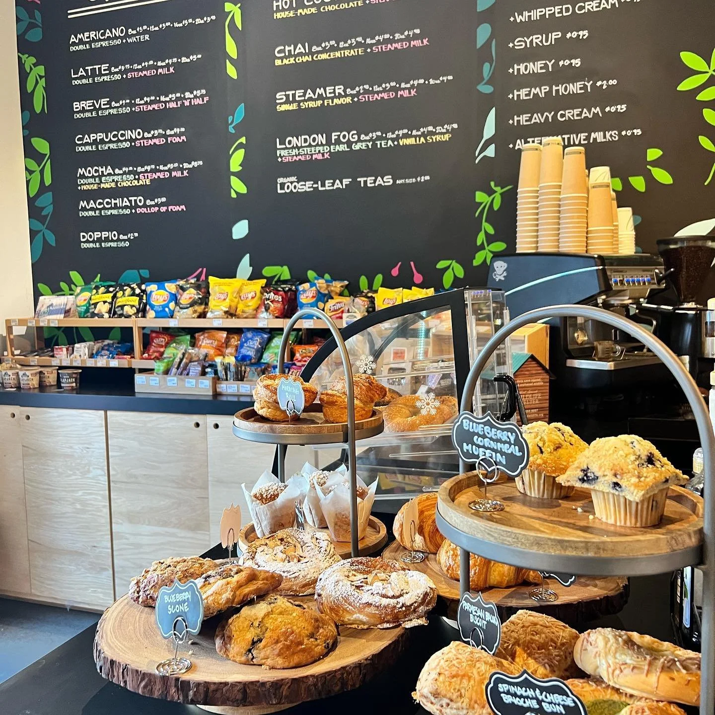 Display of various baked goods including blueberry scones, blueberry breakfast muffin, and spinach cheese brioche bun on wooden trays. In the background, shelves with snacks and a coffee menu on a chalkboard wall.