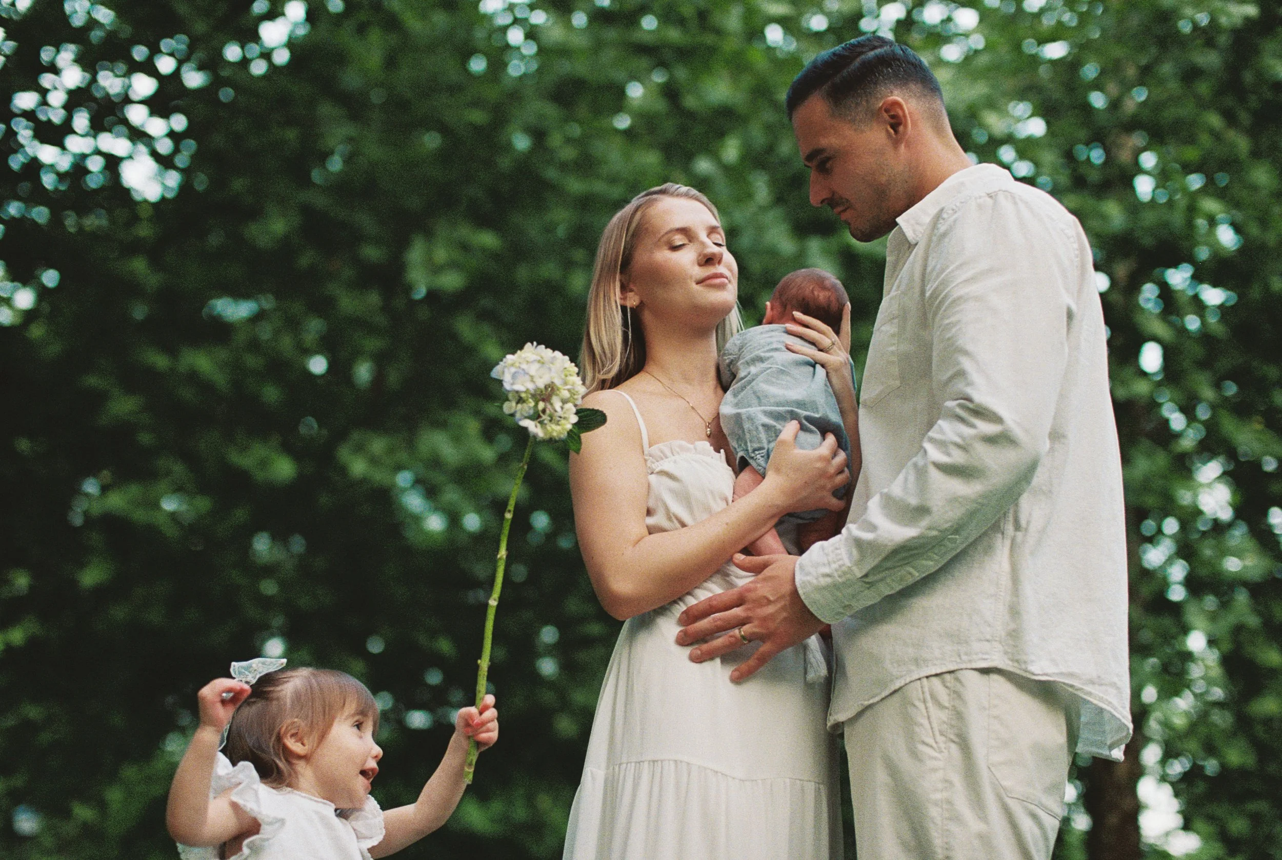Family shot of husband, wife, new baby and a toddler playing in the background