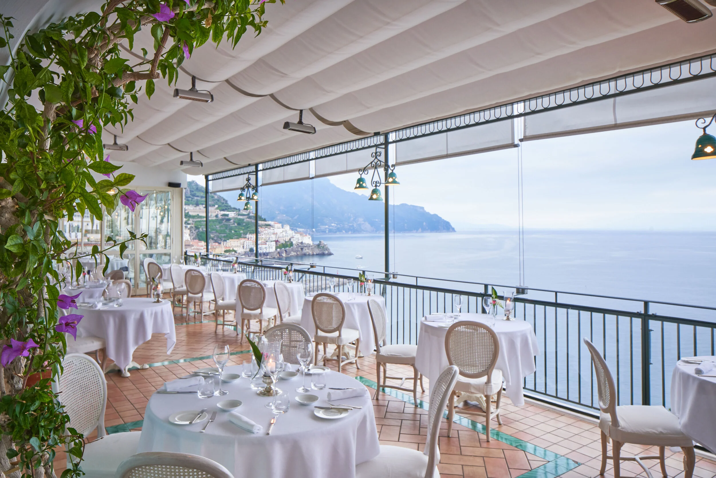Upscale outdoor restaurant patio with white tablecloths, set tables, and chairs overlooking the ocean and coastal town, under a partially covered canopy.