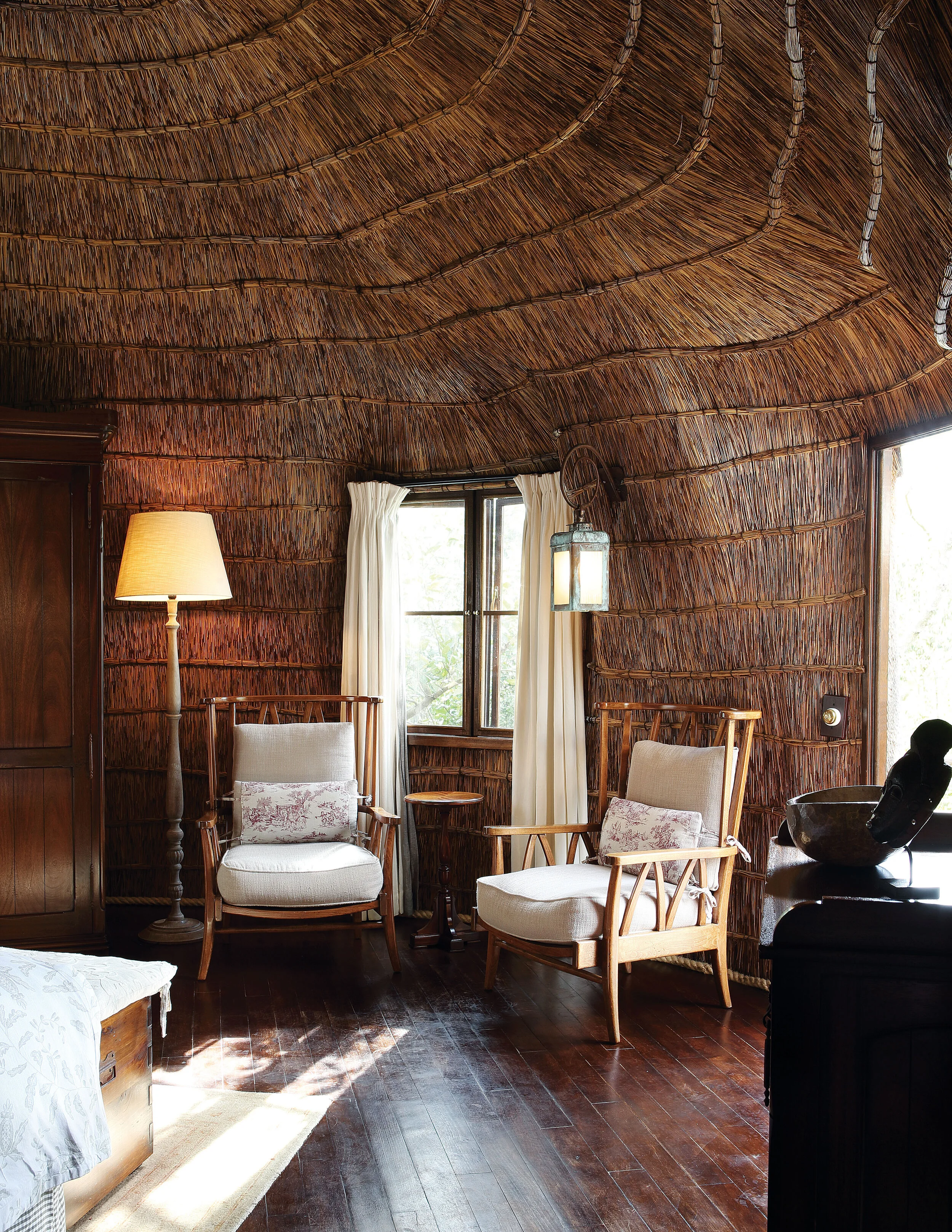 A rustic bedroom with bamboo walls and ceiling, two wooden armchairs with cushions, a standing floor lamp, a lantern hanging from the ceiling, a window with white curtains, and a wooden dresser.