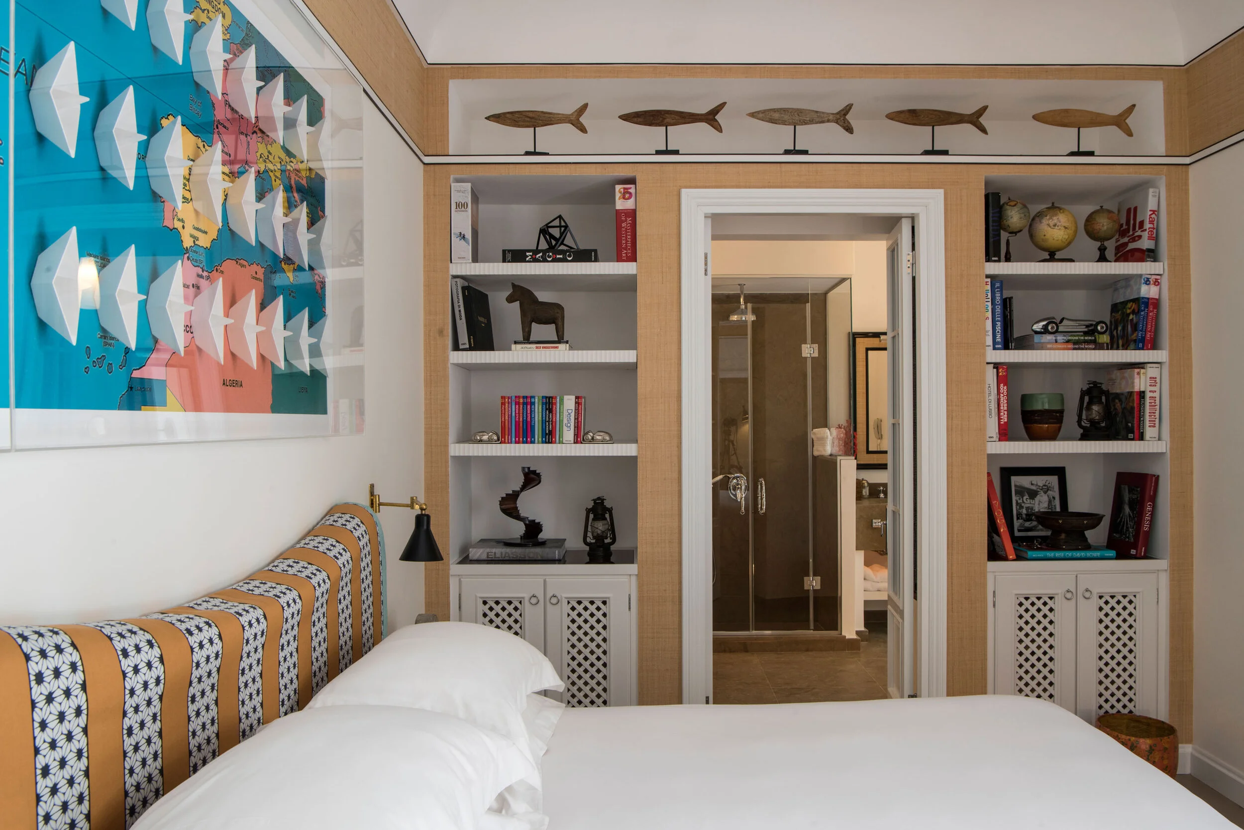 A bedroom with a bed, striped headboard, and wall-mounted lamp. Two built-in white bookshelves with various books, sculptures, globes, and decorative items on either side of a door leading to a bathroom with a glass shower.