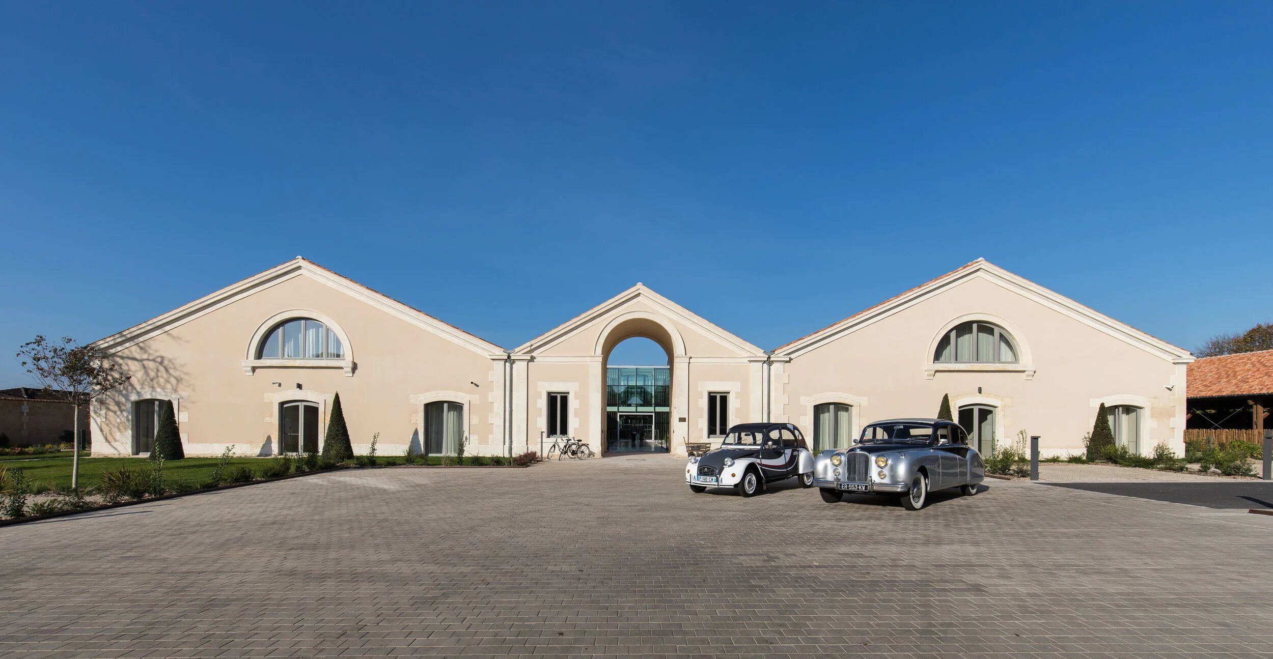 A modern building with a symmetrical design and arched windows, featuring outdoor vintage cars parked in front.