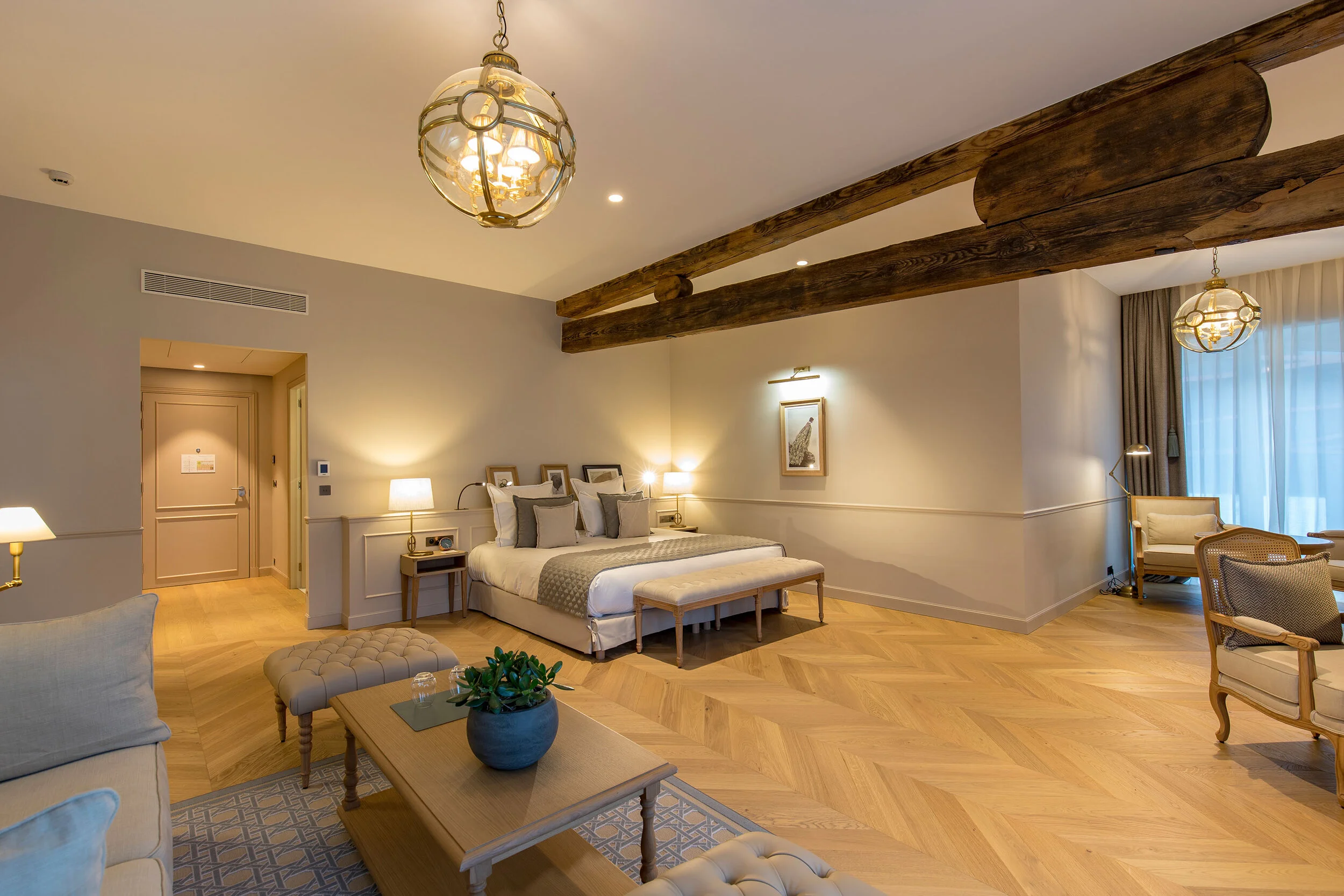 A spacious hotel room with a king-sized bed, wooden beams on the ceiling, and modern lighting and furniture, including a sitting area with a beige sofa and a wooden coffee table.