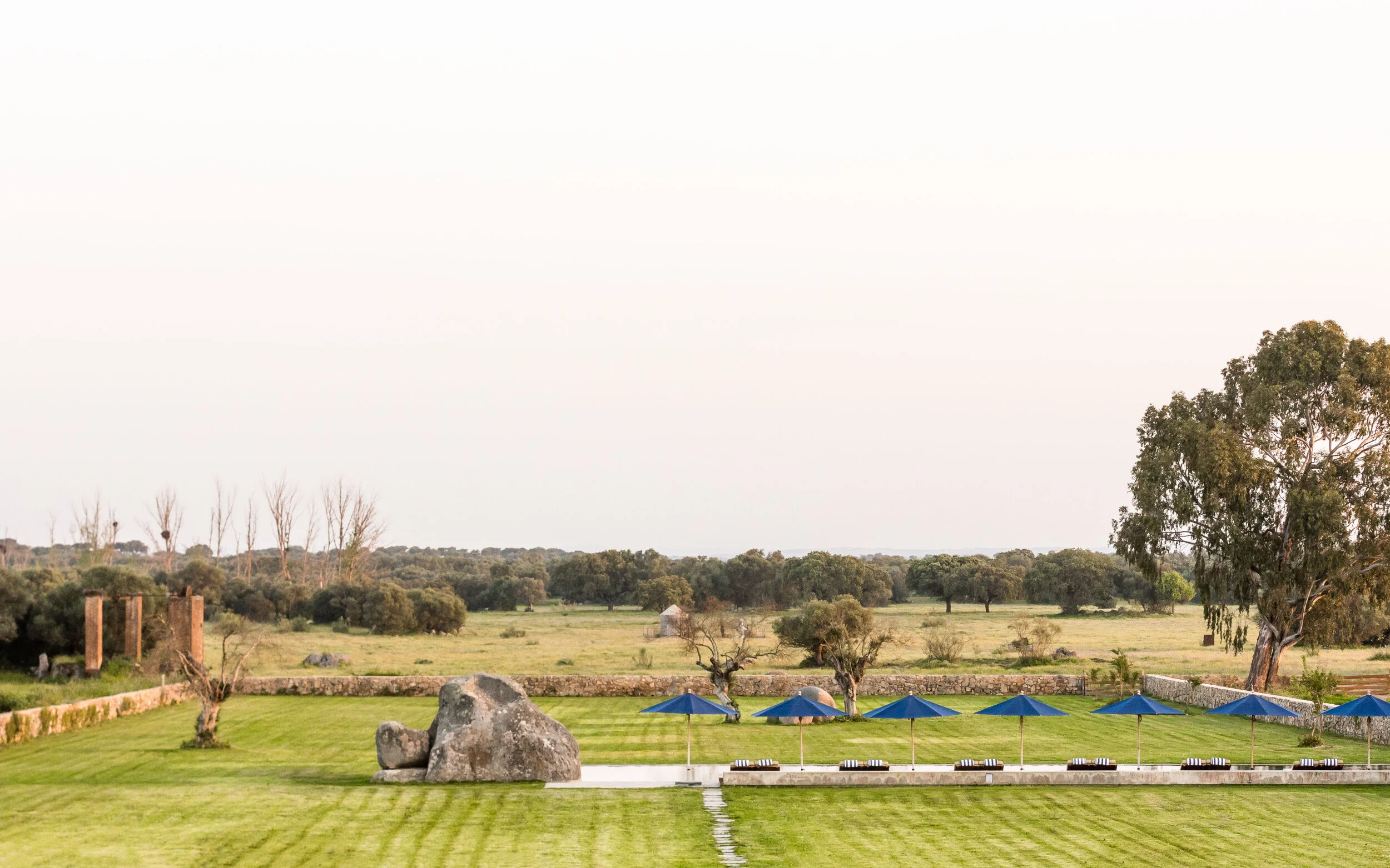 Well-maintained grassy outdoor area with large rocks, blue umbrellas, and lounge chairs, surrounded by a rural landscape with trees.