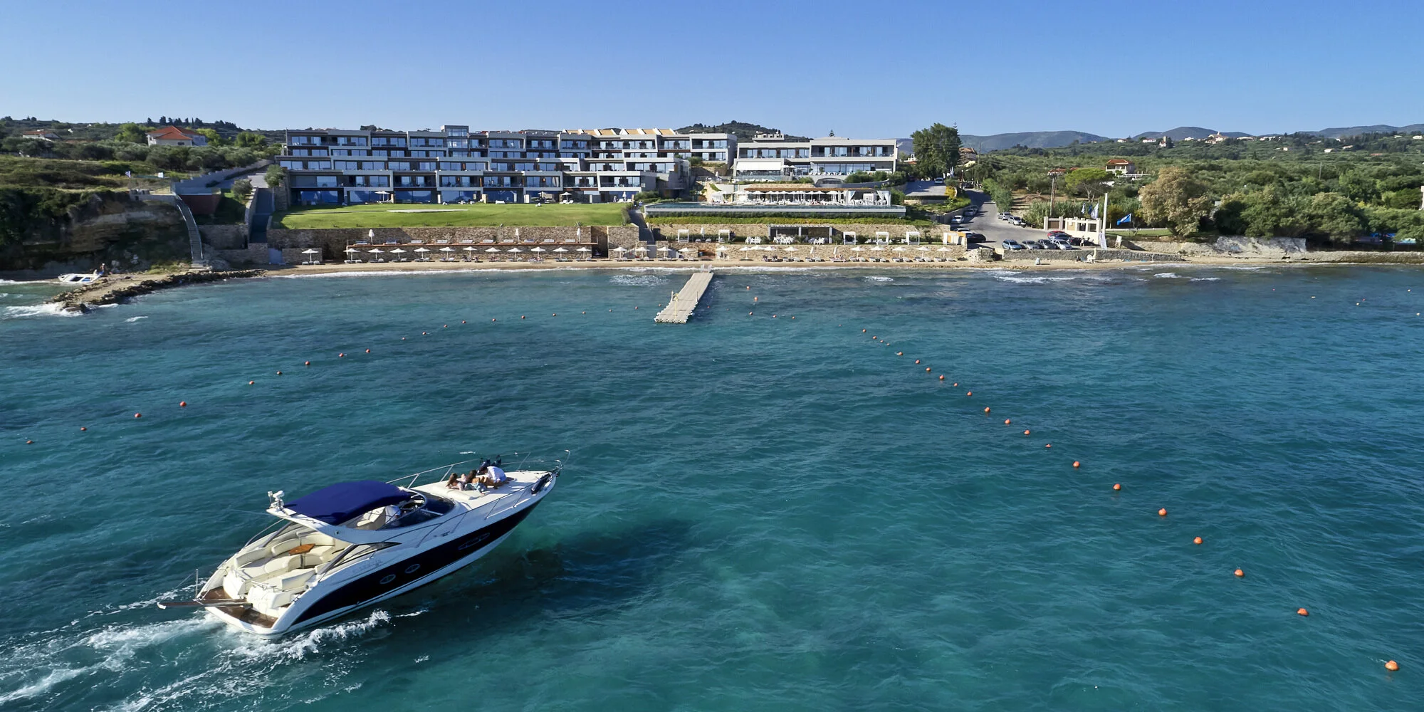 A yacht sailing in clear blue water near a coastal resort with white modern buildings and a beach with umbrellas.