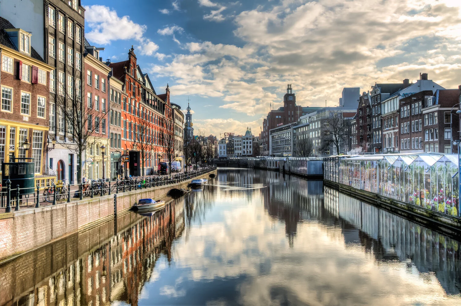 A canal lined with colorful European-style buildings and a clear sky with scattered clouds, with boats reflecting on the water, and a greenhouse with flowers on the right.