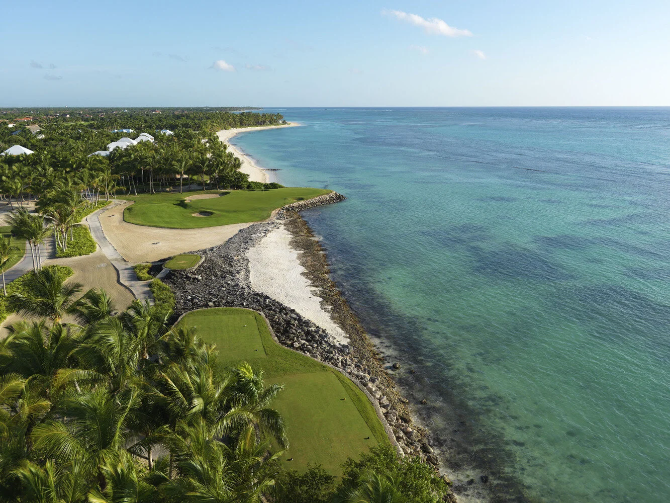 Aerial view of a coastline with a golf course, sandy beach, palm trees, and the ocean extending into the horizon.