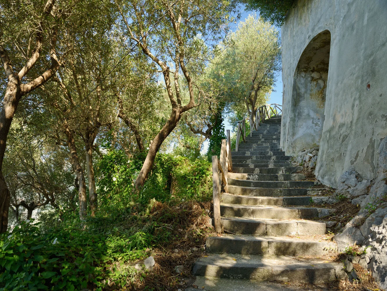 Stone stairs leading up alongside a weathered building with an arched entryway, surrounded by lush greenery and trees under a clear blue sky.