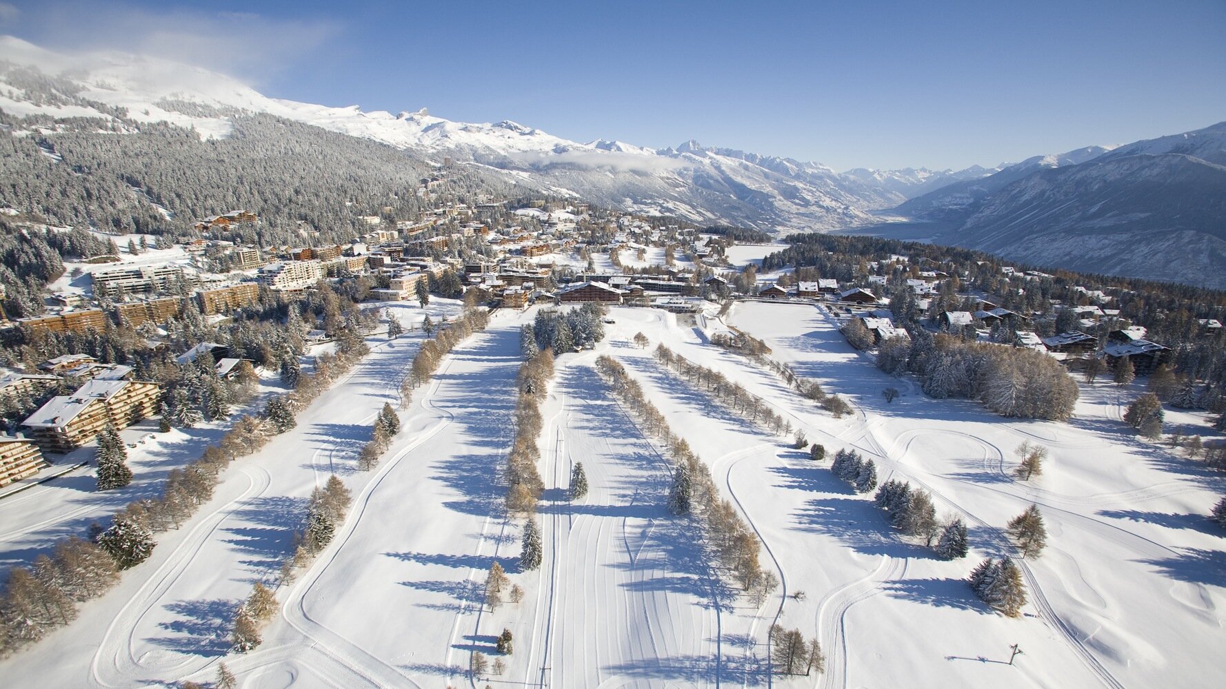 A snow-covered town in a mountainous region with ski tracks and trees in the foreground, and a mountain range in the background.