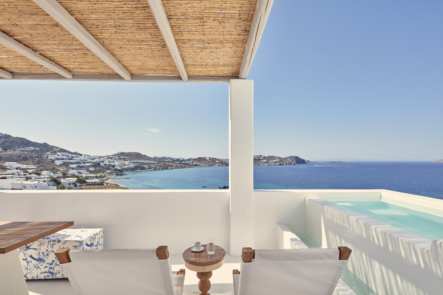 A scenic view of a coastal town seen from a balcony with white walls and modern outdoor furniture, overlooking the blue sea and clear sky.