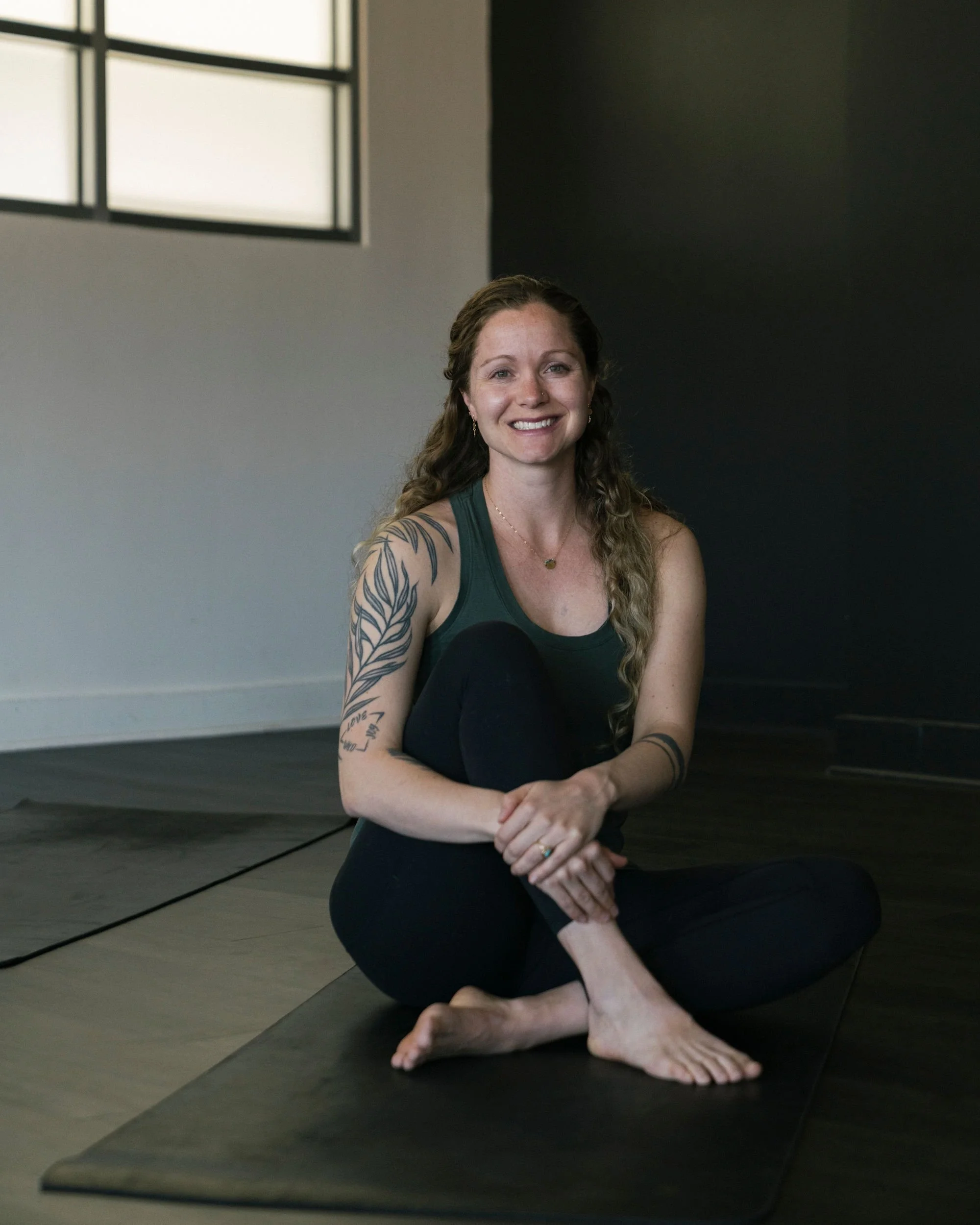 A woman with long wavy hair, wearing a dark tank top and black leggings, sitting cross-legged on a yoga mat in a room with large windows and a dark accent wall, smiling at the camera.