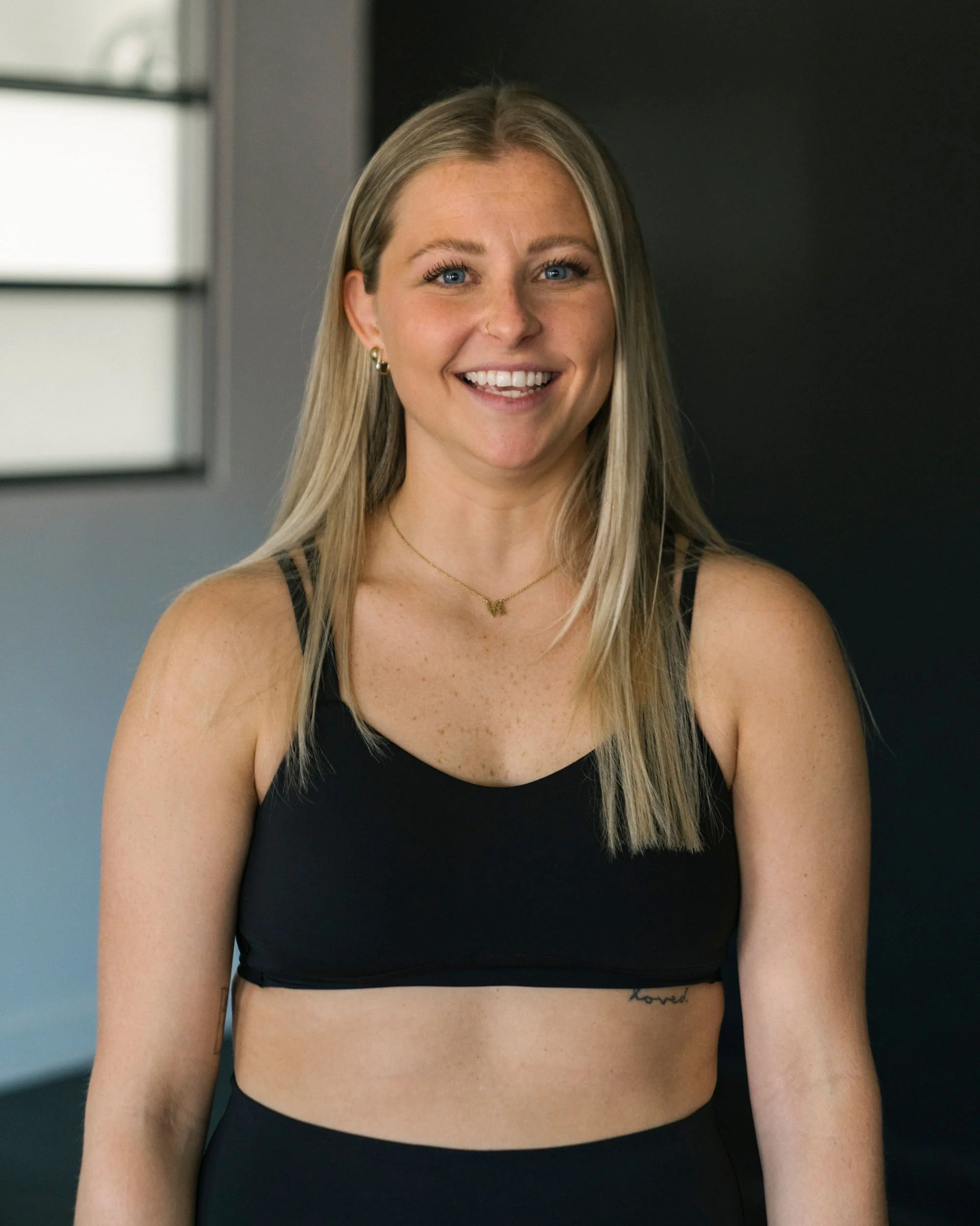 A young woman with blonde hair, blue eyes, and freckles smiling, wearing black workout clothes in a gym.
