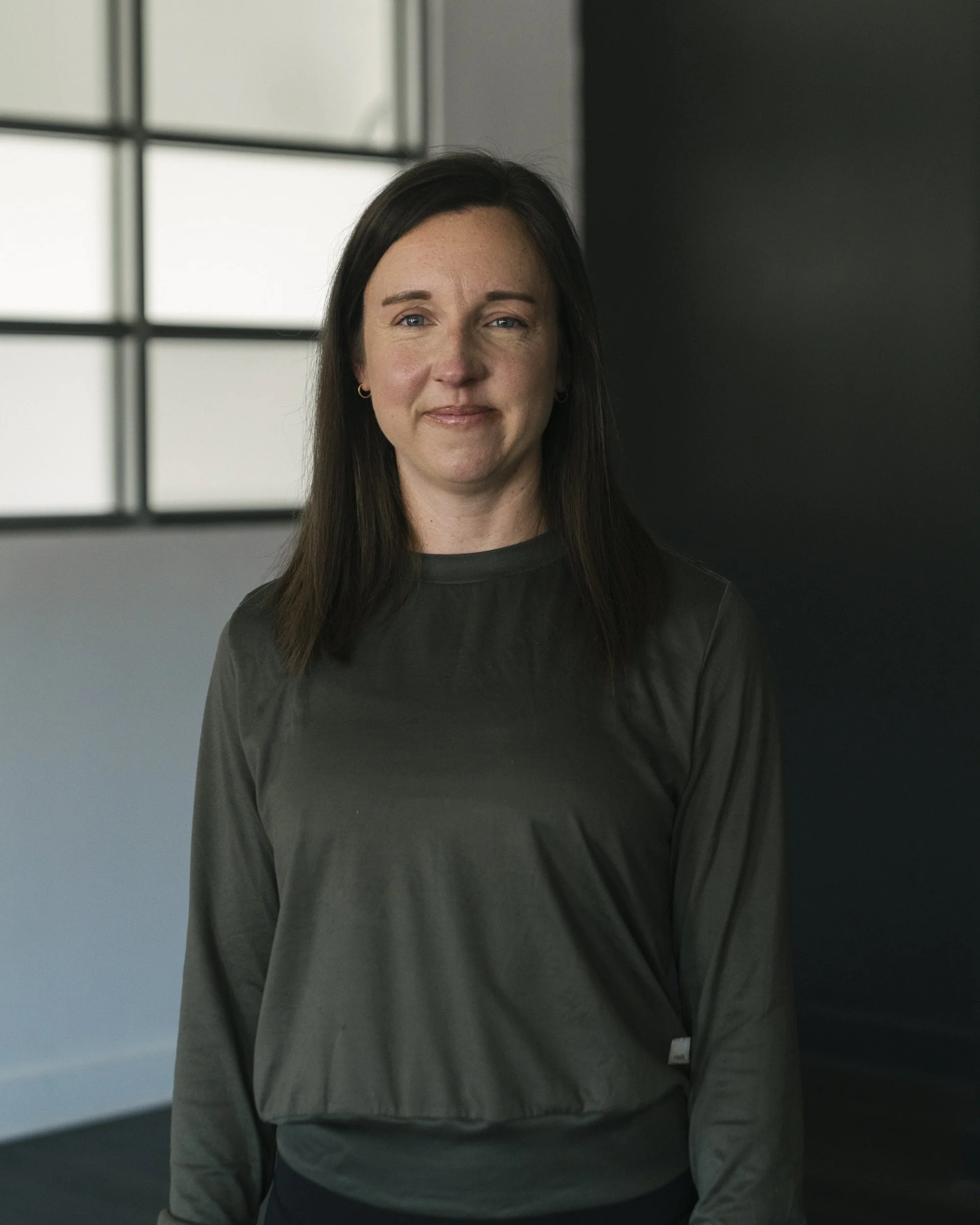A woman with dark brown hair, wearing a gray long-sleeve shirt, standing indoors near a window with black grid framing.