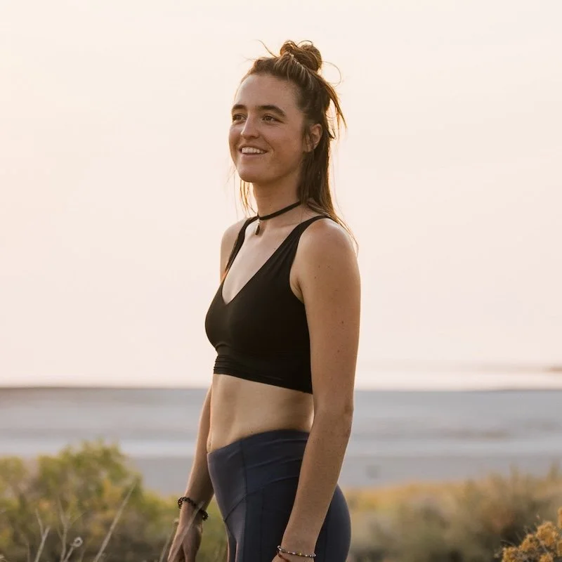 Young woman smiling outdoors near a body of water, wearing a black sports bra and high-waisted leggings.
