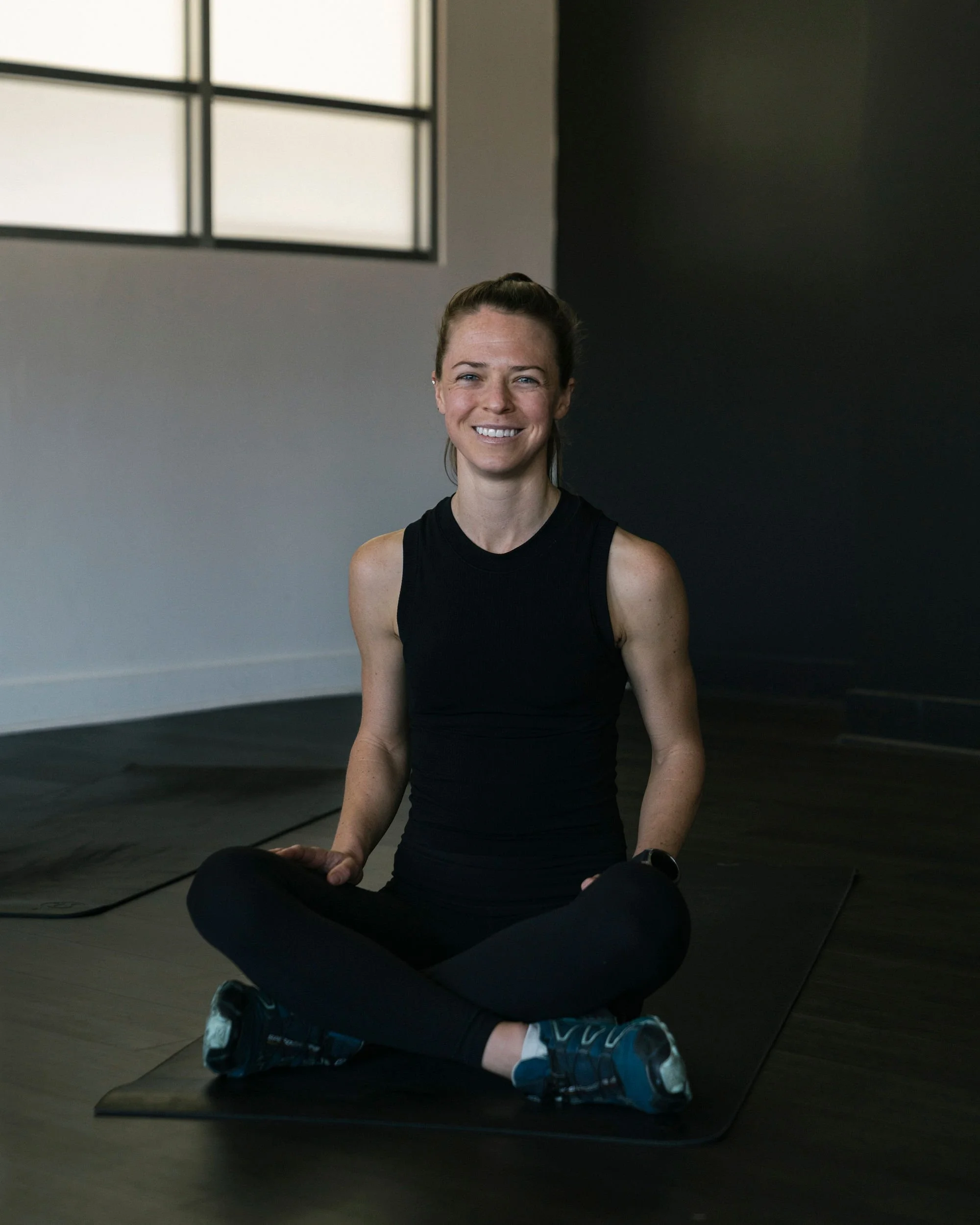 A woman sitting cross-legged on a yoga mat, smiling, in a gym or studio with large windows and dark walls.
