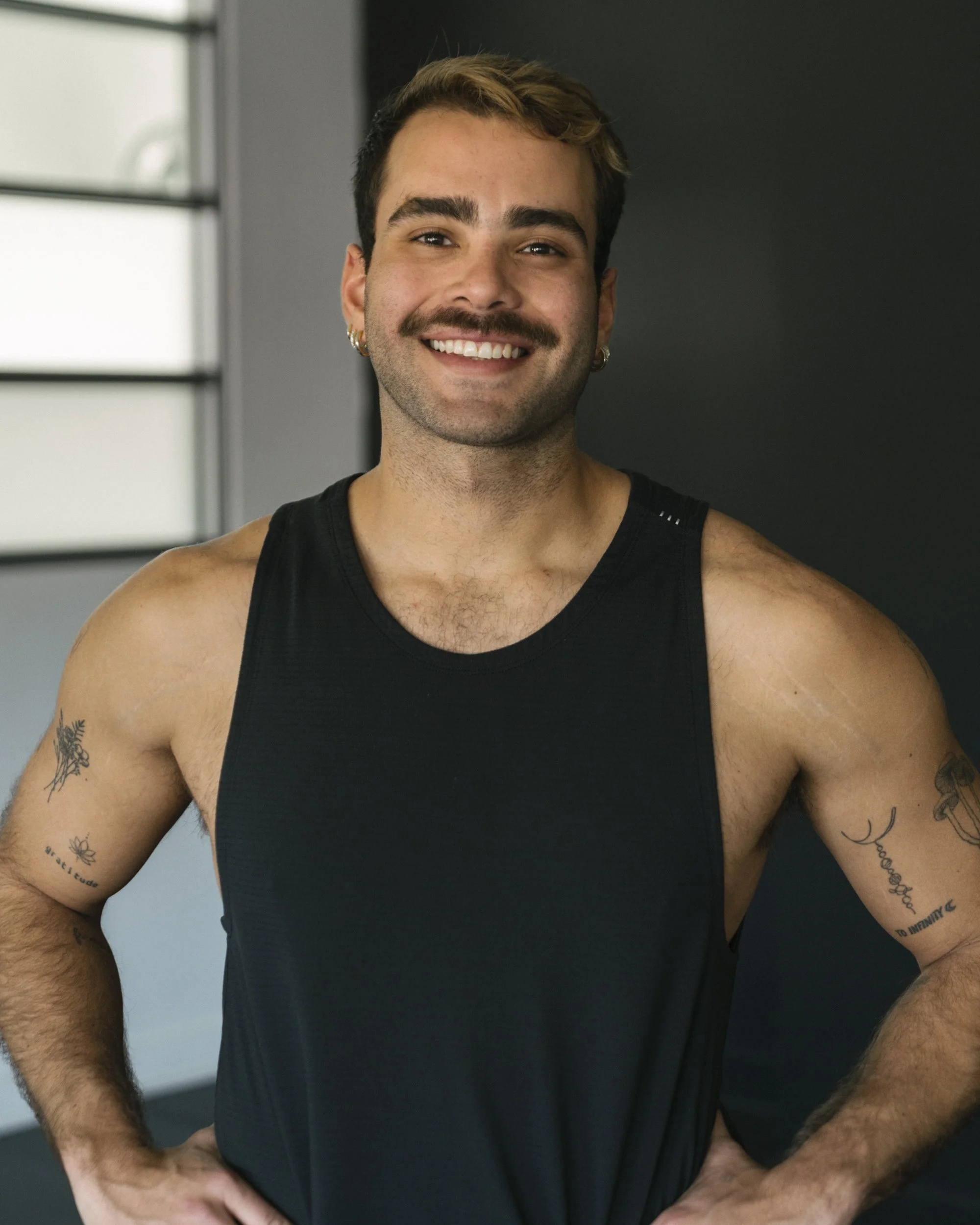 A smiling young man with a mustache and short, styled hair, wearing a black sleeveless athletic shirt, standing indoors near a window with blinds.