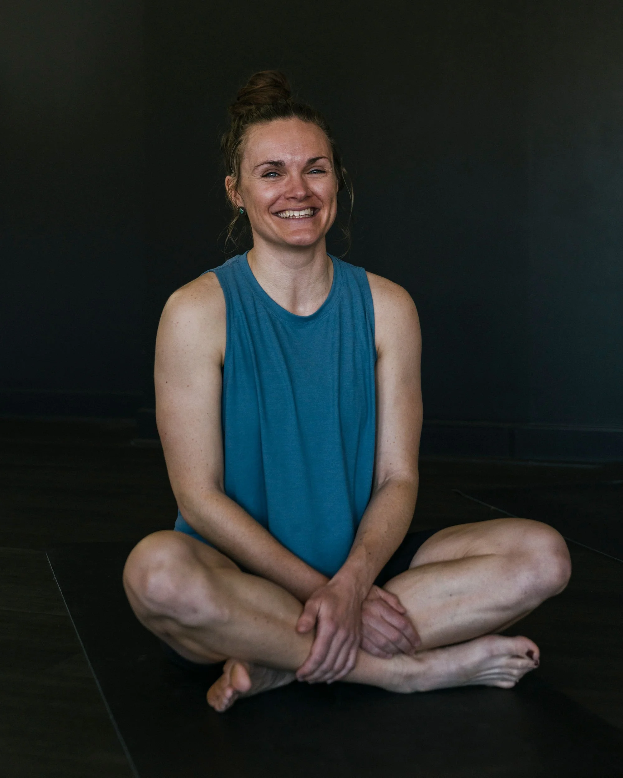 A woman sitting cross-legged on a floor, wearing a blue sleeveless top and black shorts, smiling with her hair in a bun against a plain dark background.