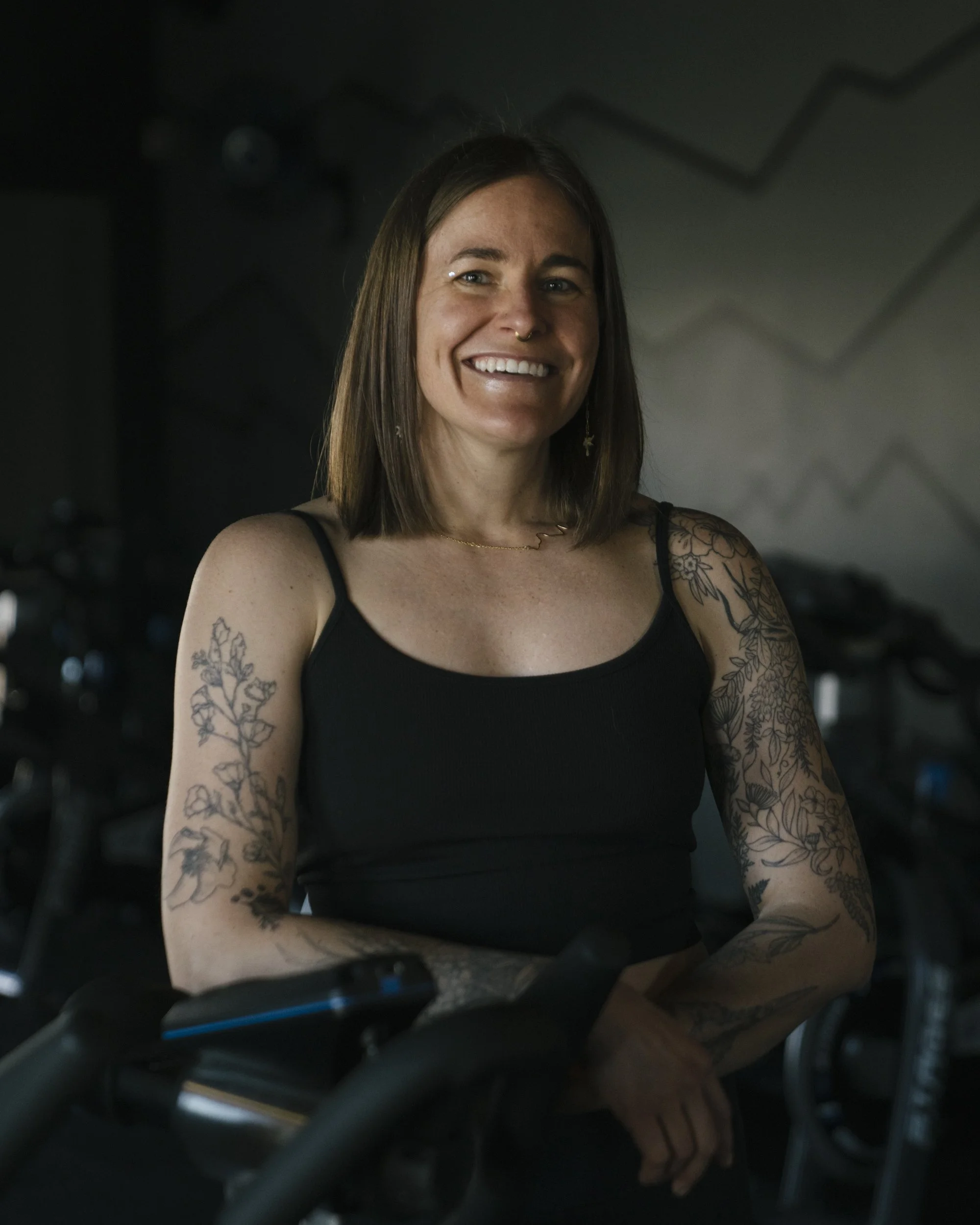 A smiling woman with shoulder-length brown hair and tattoos on her arms, wearing a black tank top, sitting on a bicycle in a gym.