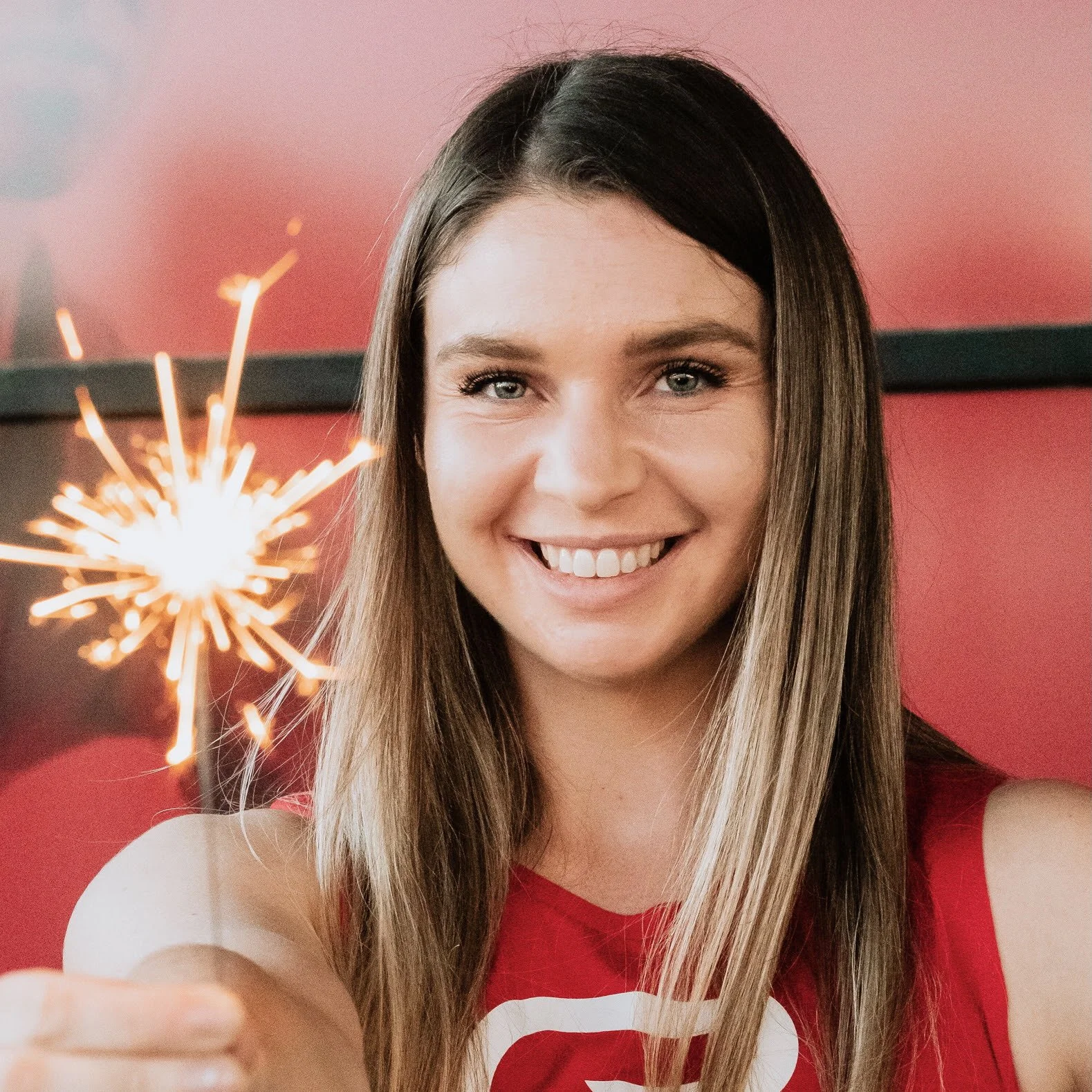A young woman with long brown hair and blue eyes smiling while holding a lit sparkler in front of a red background.