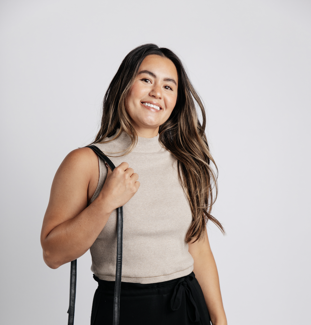 A woman with long brown hair, wearing a sleeveless beige top and black pants, smiling while holding a backpack strap over her shoulder against a plain gray background.