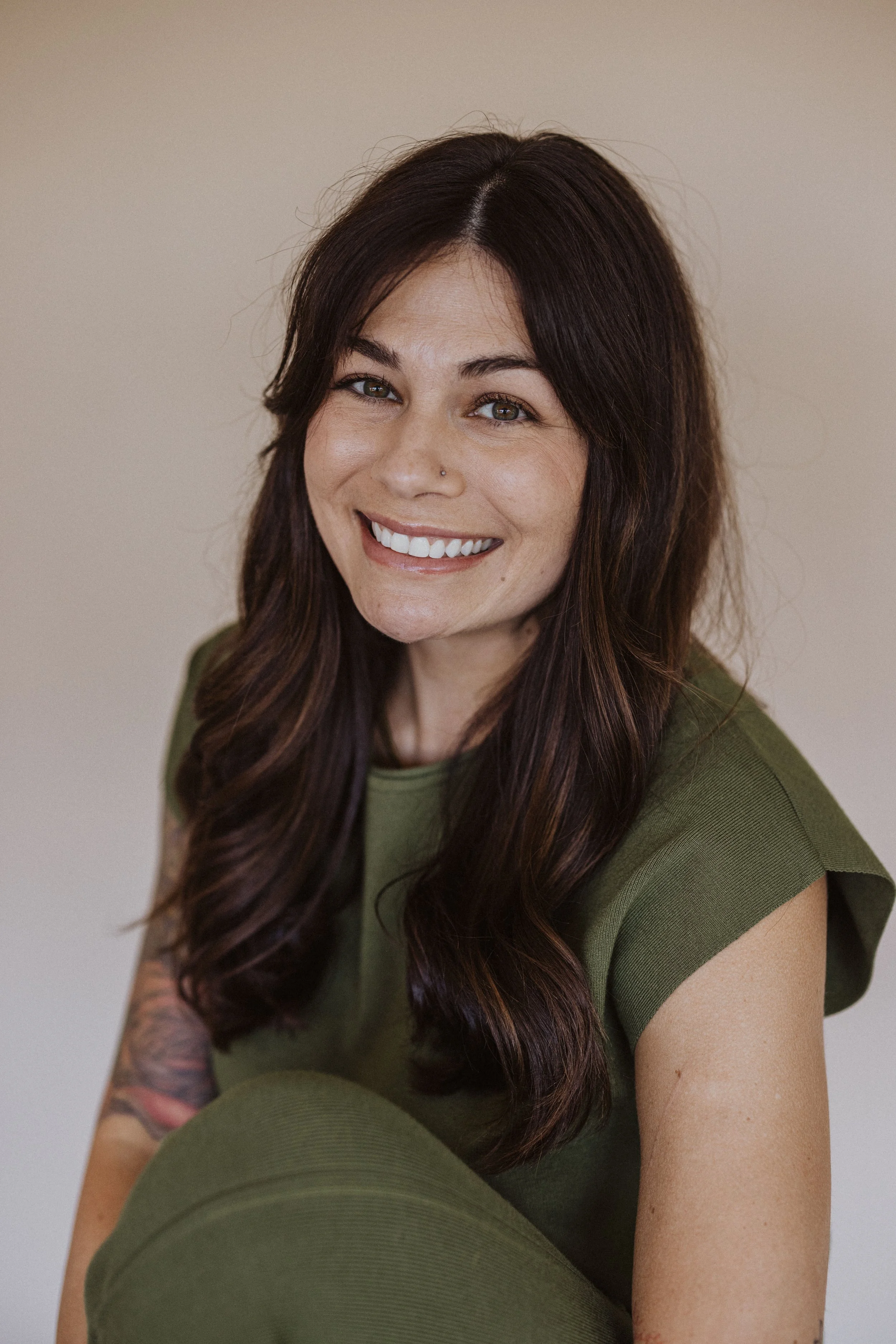 A woman with long, dark brown hair, smiling, wearing an olive green top, with a tattoo on her right arm, sitting against a beige background.