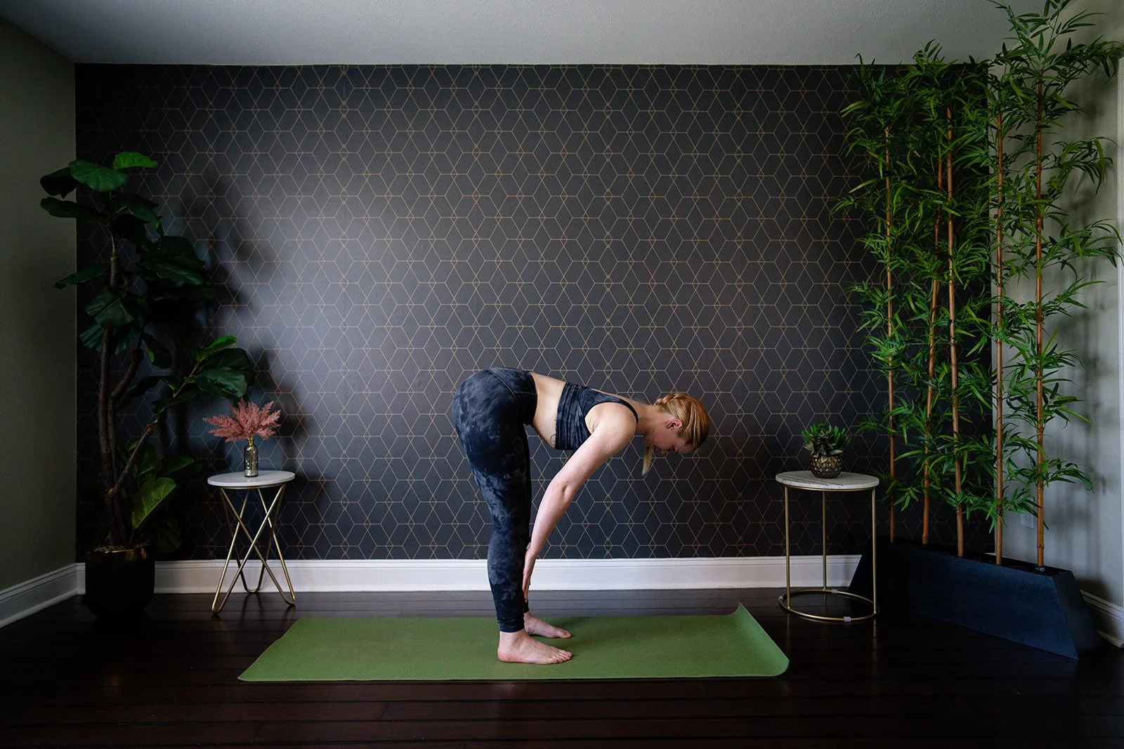 Woman practicing yoga forward bend pose on mat in stylish room with decorative plants and patterned wall.