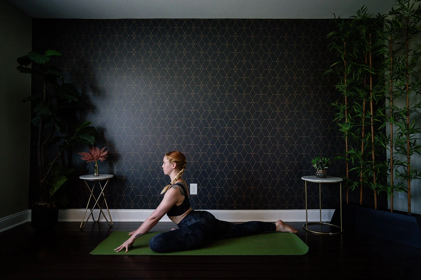 Woman practicing yoga in pigeon pose on green mat indoors with modern decor and plants.